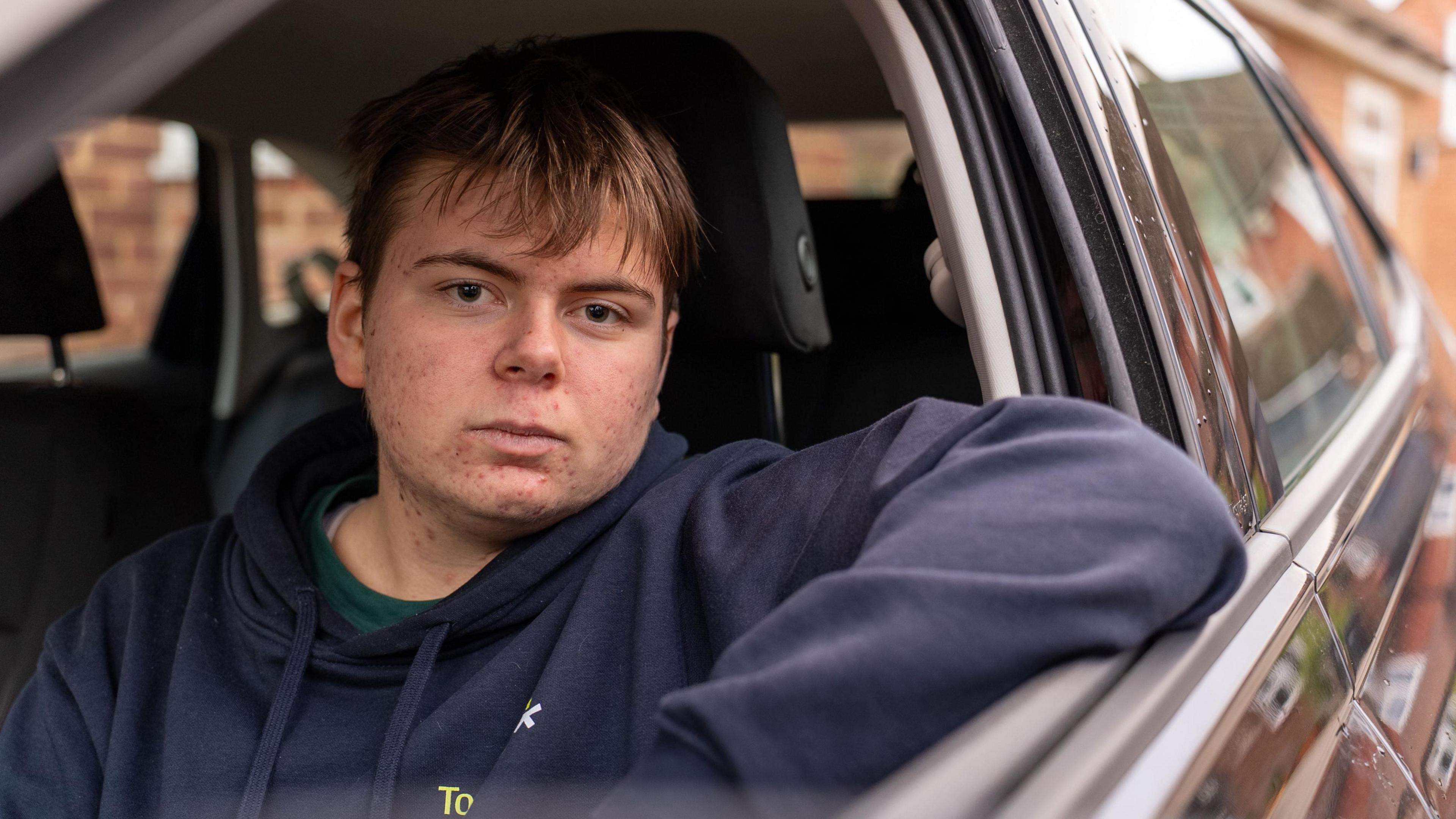 Young 18 year old man sits in the passenger seat of a black VW car, his arm resting on the open window as he looks to camera
