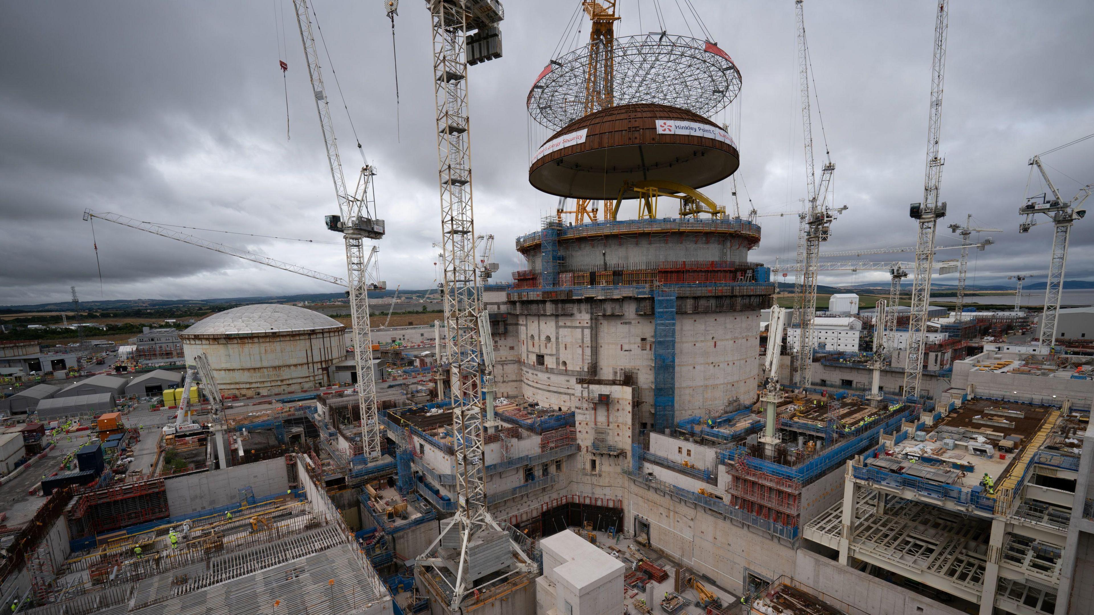 The nuclear power station construction site at Hinkley Point C is seen in a wide shot showing multiple buildings under construction and cranes. In the centre of the image a large dome is being lifted onto a cylinder-shaped building
