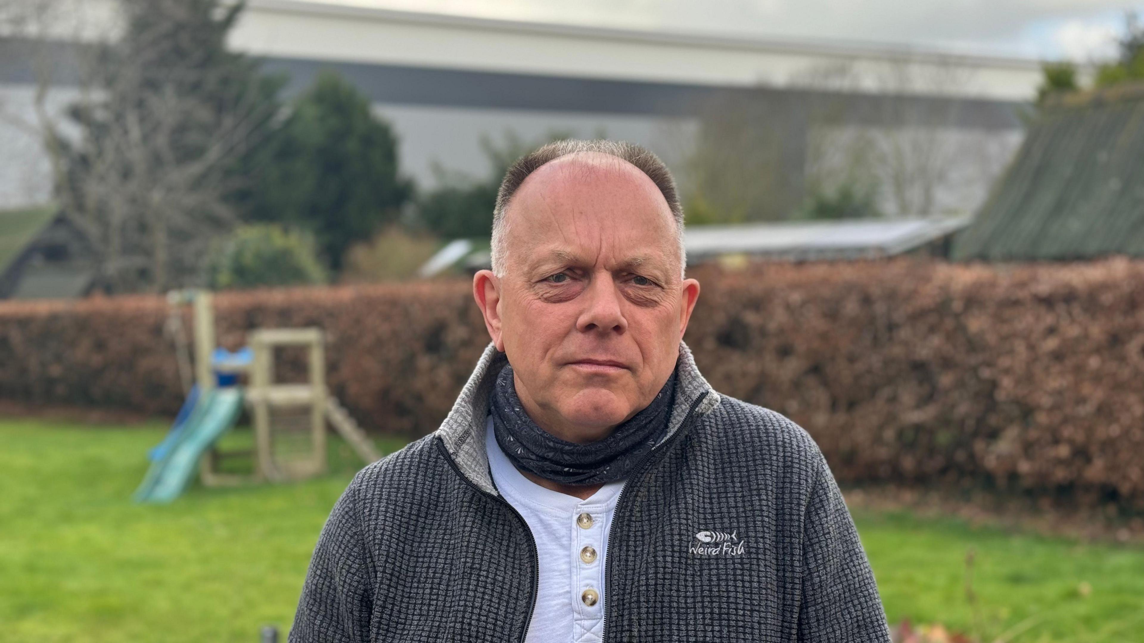 Adrian Day is wearing a grey jumper over a white top and a scarf. He is standing in a garden and the large warehouse can be seen in the background.