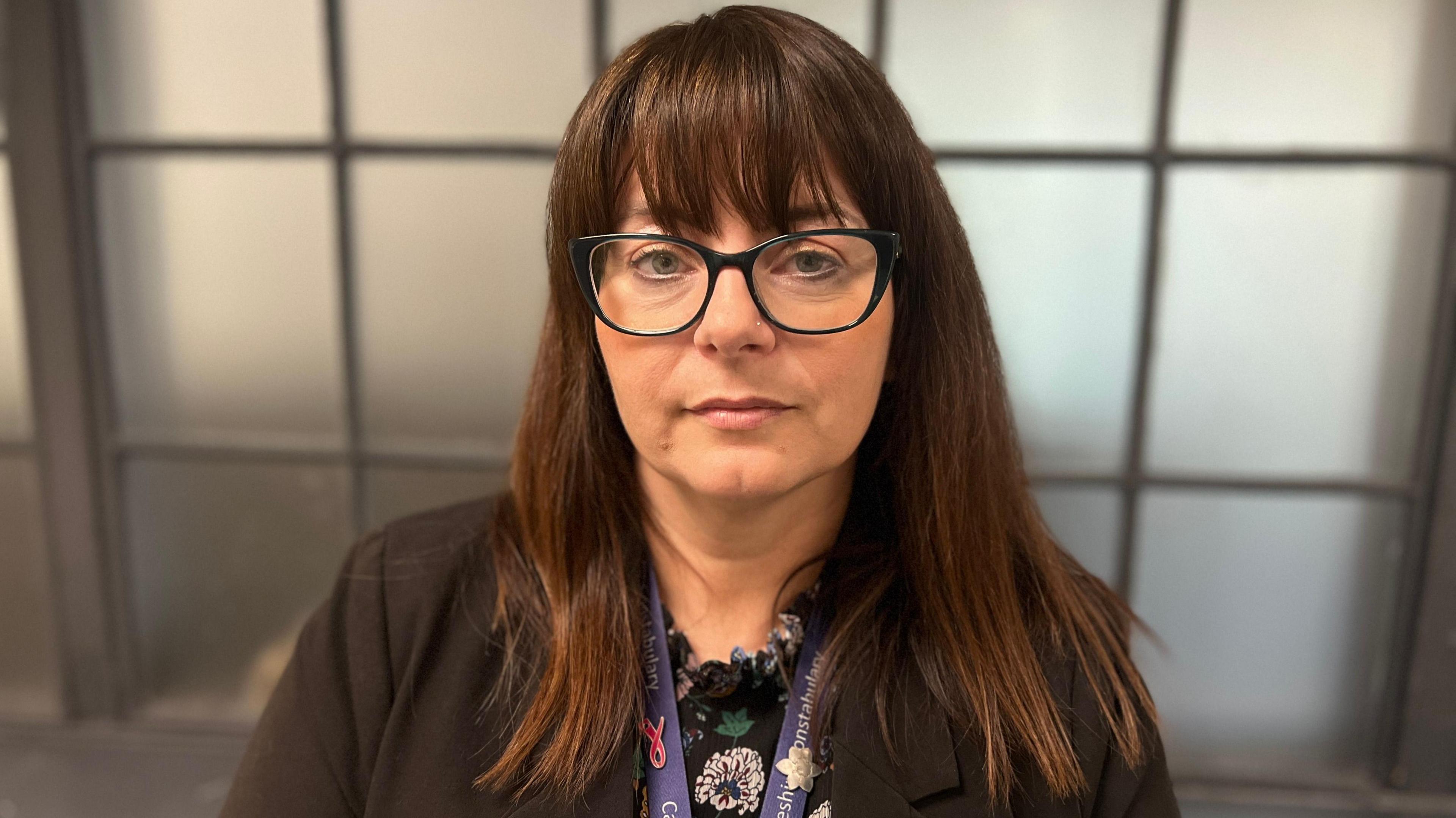 Annette Renwick is outside an translucent window wearing a dark jacket and ID lanyard. She has long brown hair, a fringe and is wearing glasses.
