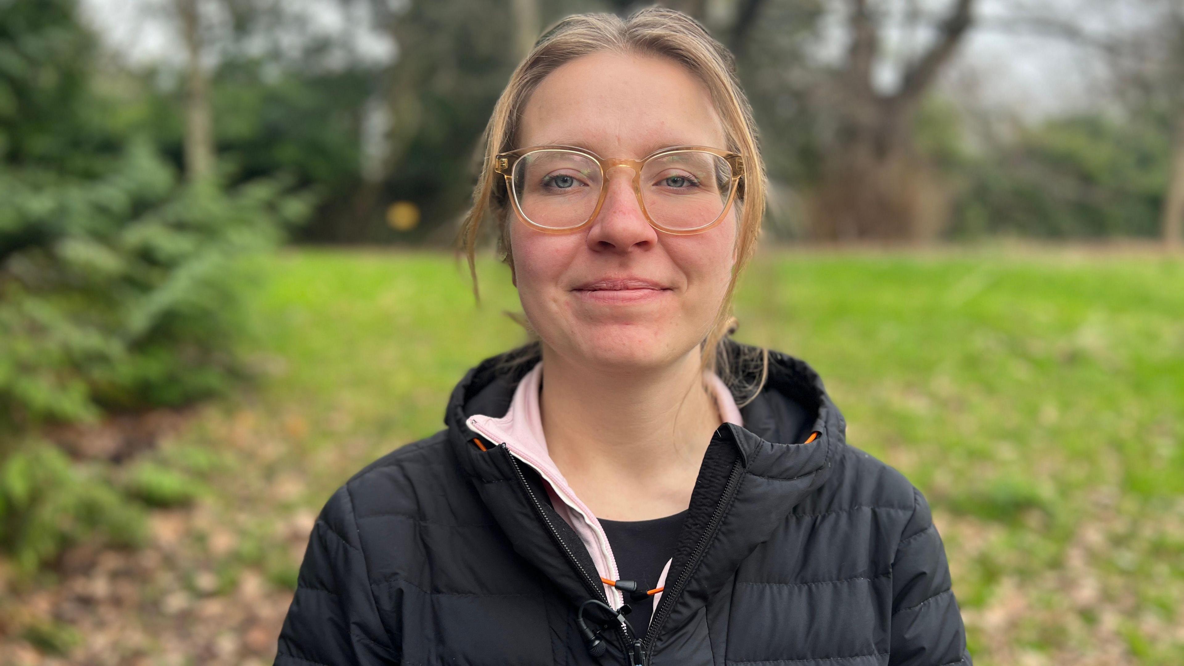 Karolina Pakenaite. She has long blonde hair which is tied up and glasses. She is wearing a black puffer coat over a pink fleece and a black t-shirt. She is pictured outside, standing in front of a field and some trees. She is looking directly at the camera and smiling. 