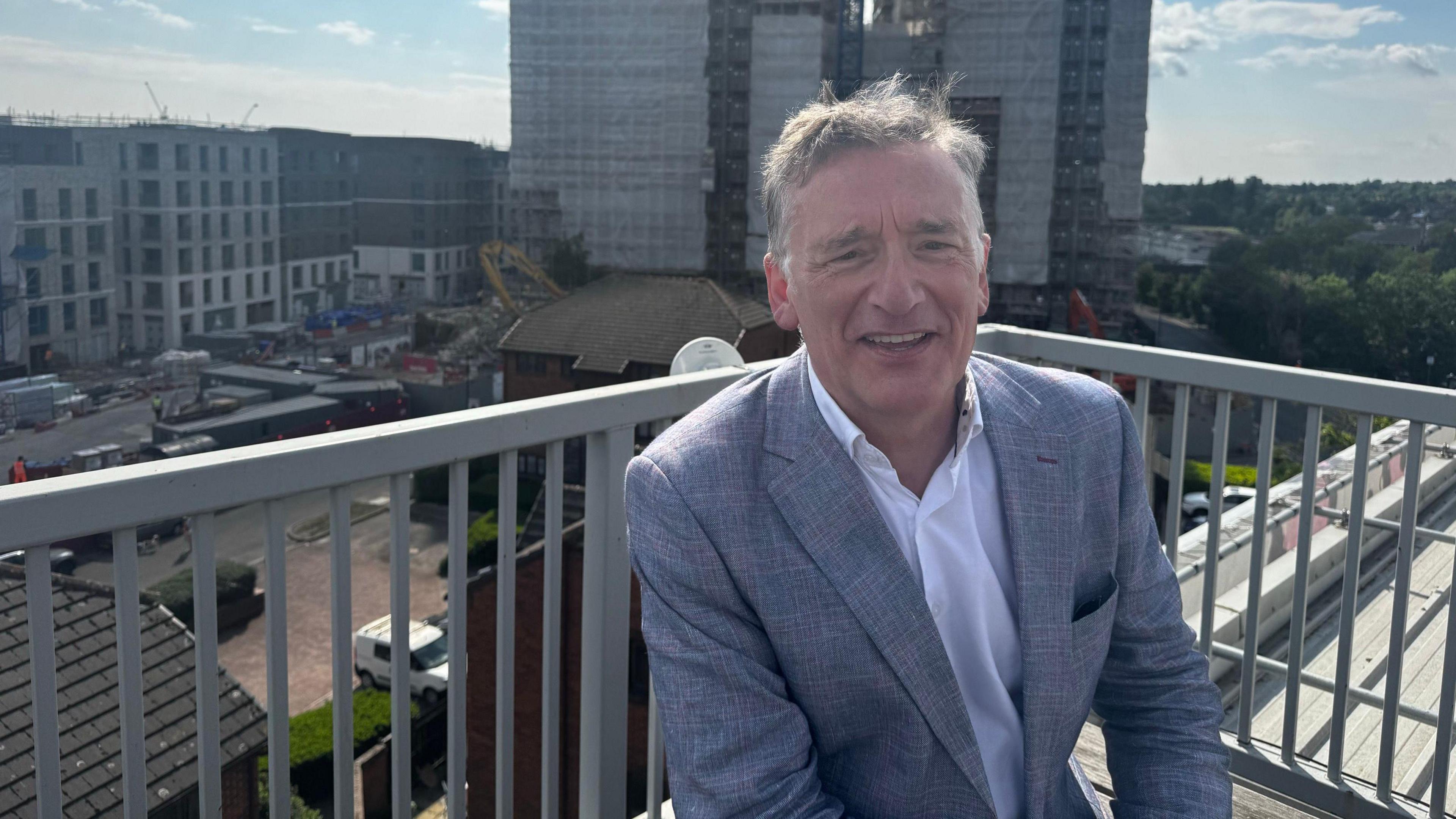 Looking towards the camera wearing a white shirt and navy suit, Ian McDermott smiles against the backdrop of a grey balcony and a construction site somewhere in London