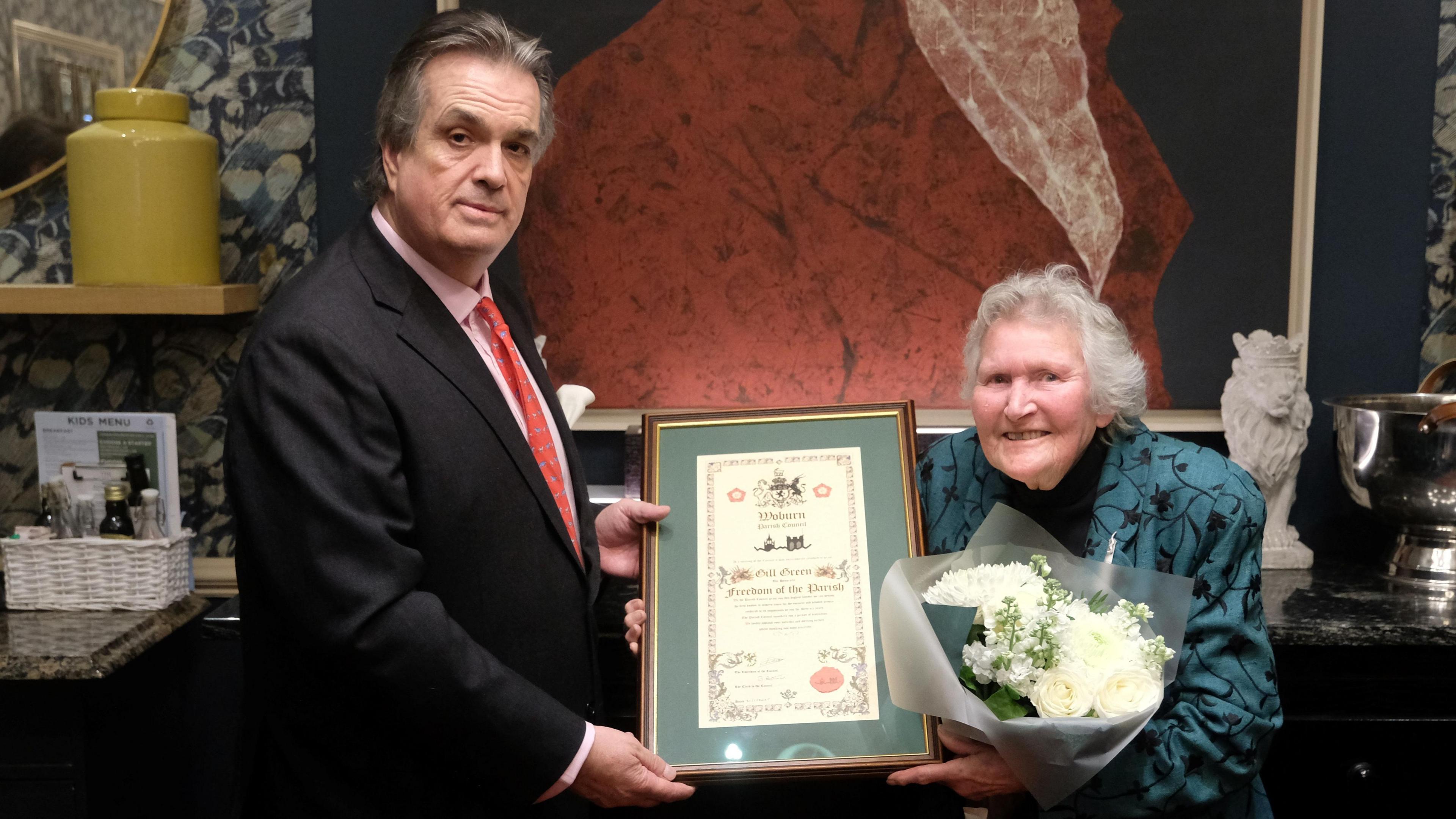 Gill Green (left) holding a bouquet of white flowers and a certificate in a frame. She is wearing a green and black jacket. The Duke of Bedford, who has short dark hair and is wearing a dark suit and red tie, is standing next to her. He is holding the other side of the certificate.