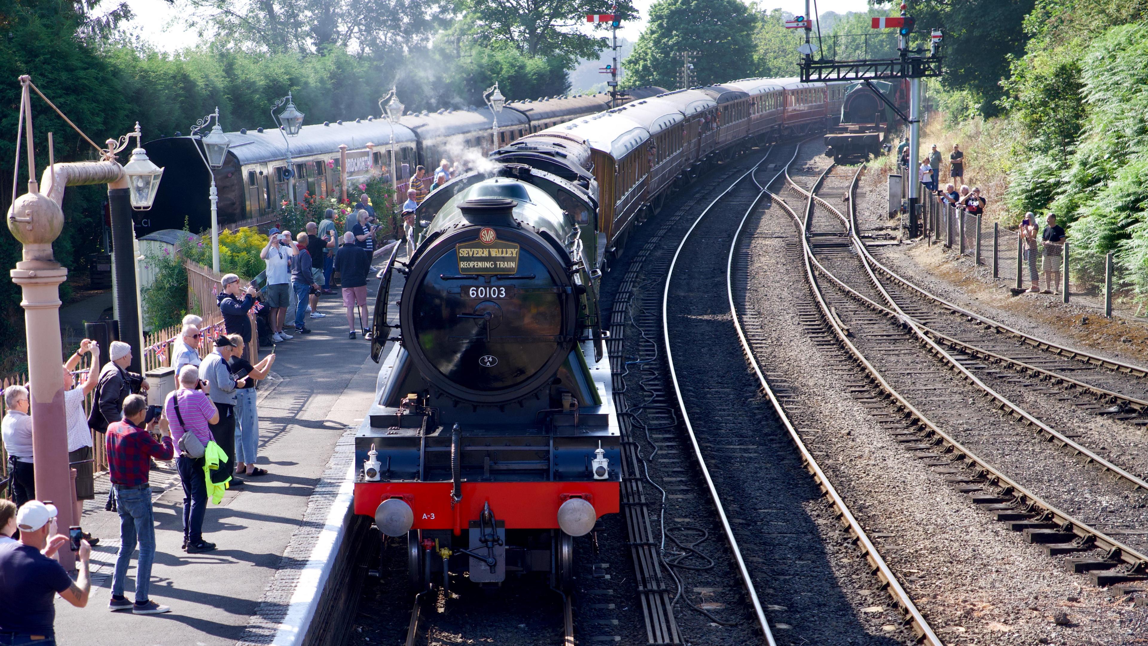 Flying Scotsman locomotive arrives at Bridgnorth. The green loco has a black boiler cover with a sign that reads Severn Valley reopening train. Behind the loco are seven brown carriages with people on the platform taking photographs with their mobile phones