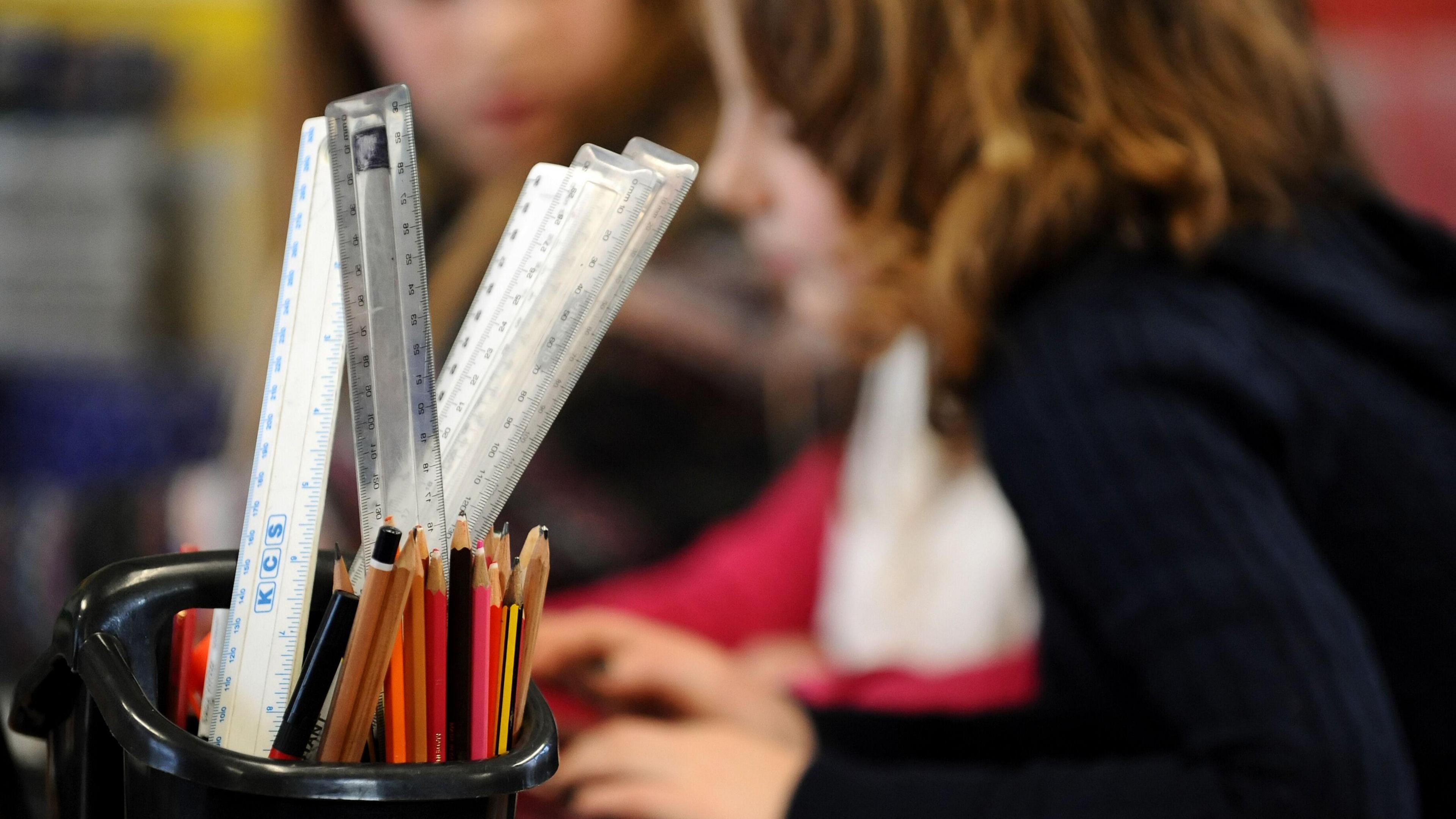 Pencil pot in foreground with pencils and rulers in. Two female primary school students in the background, the closer one has mousy brown, curly, shoulder length hair you are unable to see any identifying features of the second student.