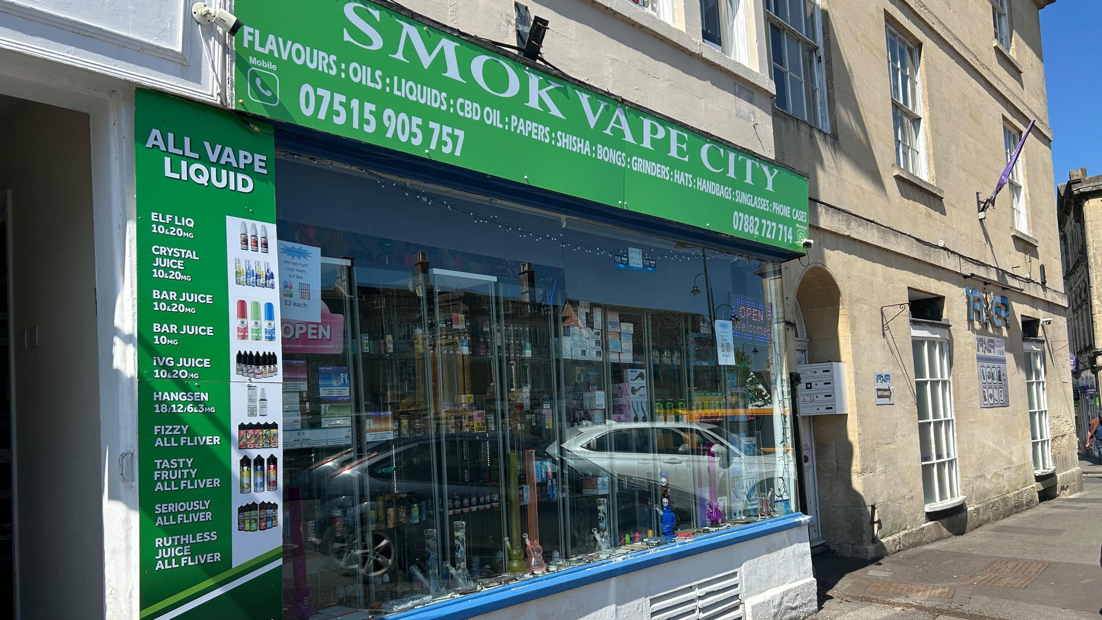 The store front of Smok Vape City in Chippenham. There is a big glass front with vapes on display. A bright green sign sits above the glass screen with the shops name and number on it. To the left, there is another green sign which lists the vape liquids which are sold.