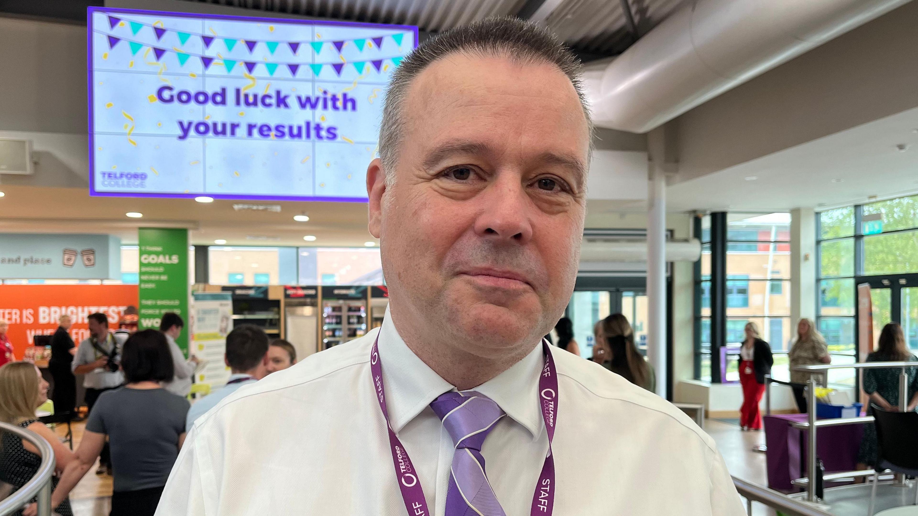 Lawrence pictured looking into the camera and smiling slightly. He's wearing a white shirt and a purple striped tie, with a purple Telford College lanyard. Behind him is a busy school atrium, which has white walls and floor-to-ceiling windows. There's a big screen hanging up on the wall behind him, which reads 'Good luck with your results' in purple letters on a white background, with a picture of blue and purple bunting and yellow confetti. 