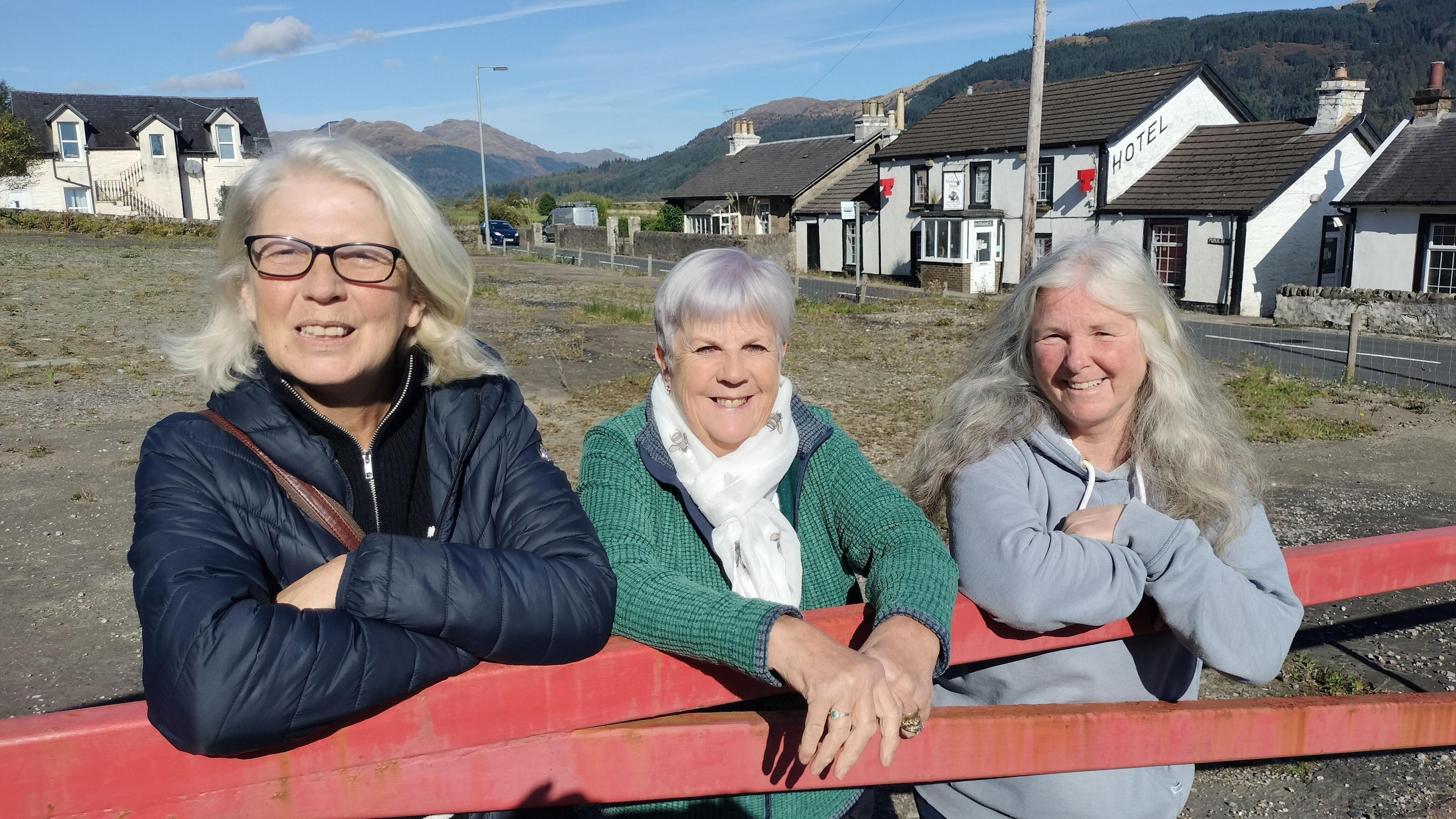 Three woman stand leaning on a red fence. The hotel is in the background, with the mountains in the distance.
Caroline stands with her arms folded and wears glasses. She has light blonde hair and smiles whilst wearing a navy puffer jacket.
Sue has short grey hair and smiles at the camera. She is wearing a green jacket and a white scarf. Her elbows are leaning on the fence with her hands slightly clasped. 
Helen smiles at the camera and has long blonde/grey hair. She has her arms folded whilst leaning on the fence and wears a grey hoodie. 