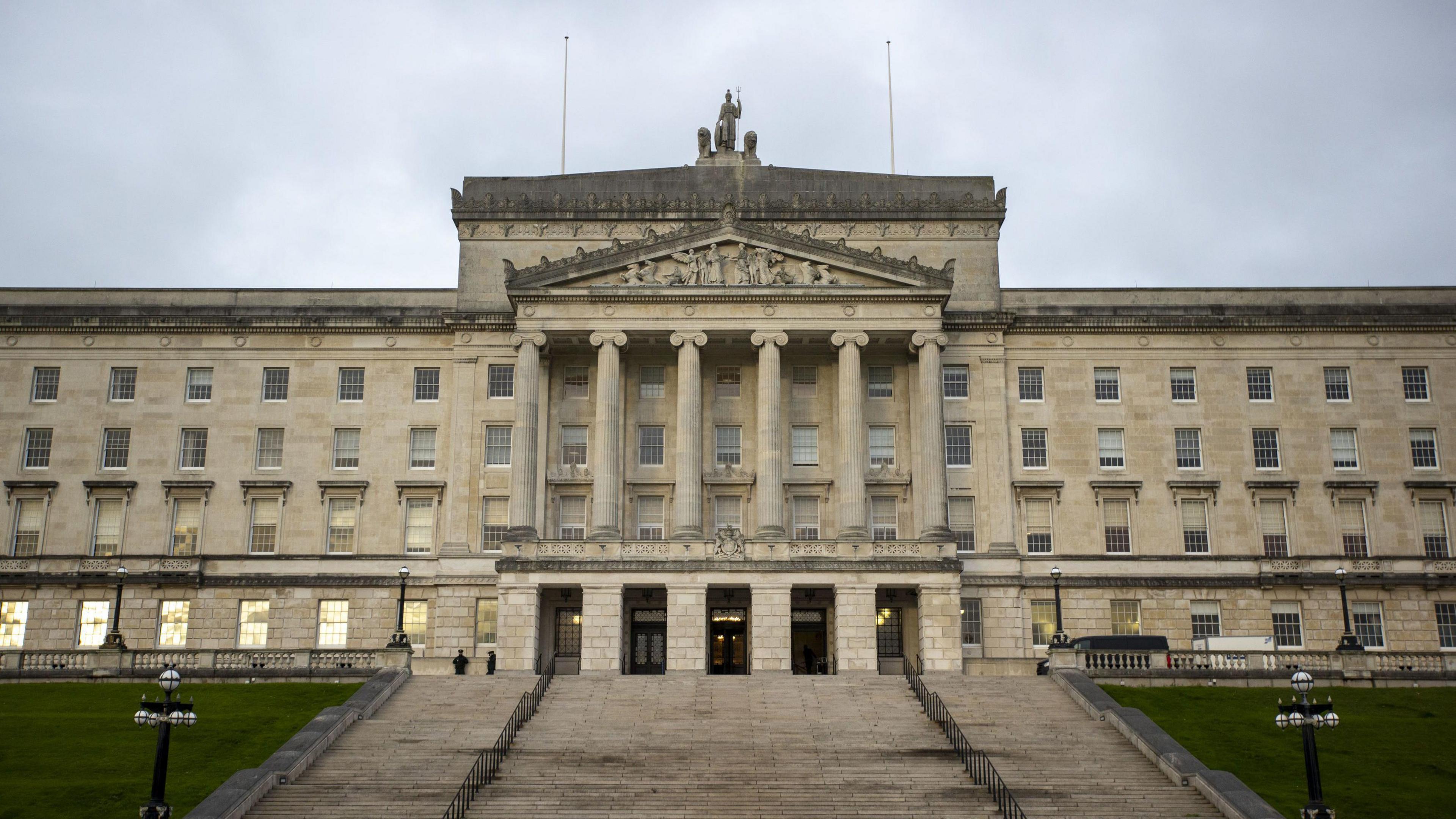 A wide shot of the exterior of Stormont/Parliament Buildings. It features a large, white-stone building with six large columns in the front with a large, wide staircase leading up to the doors.