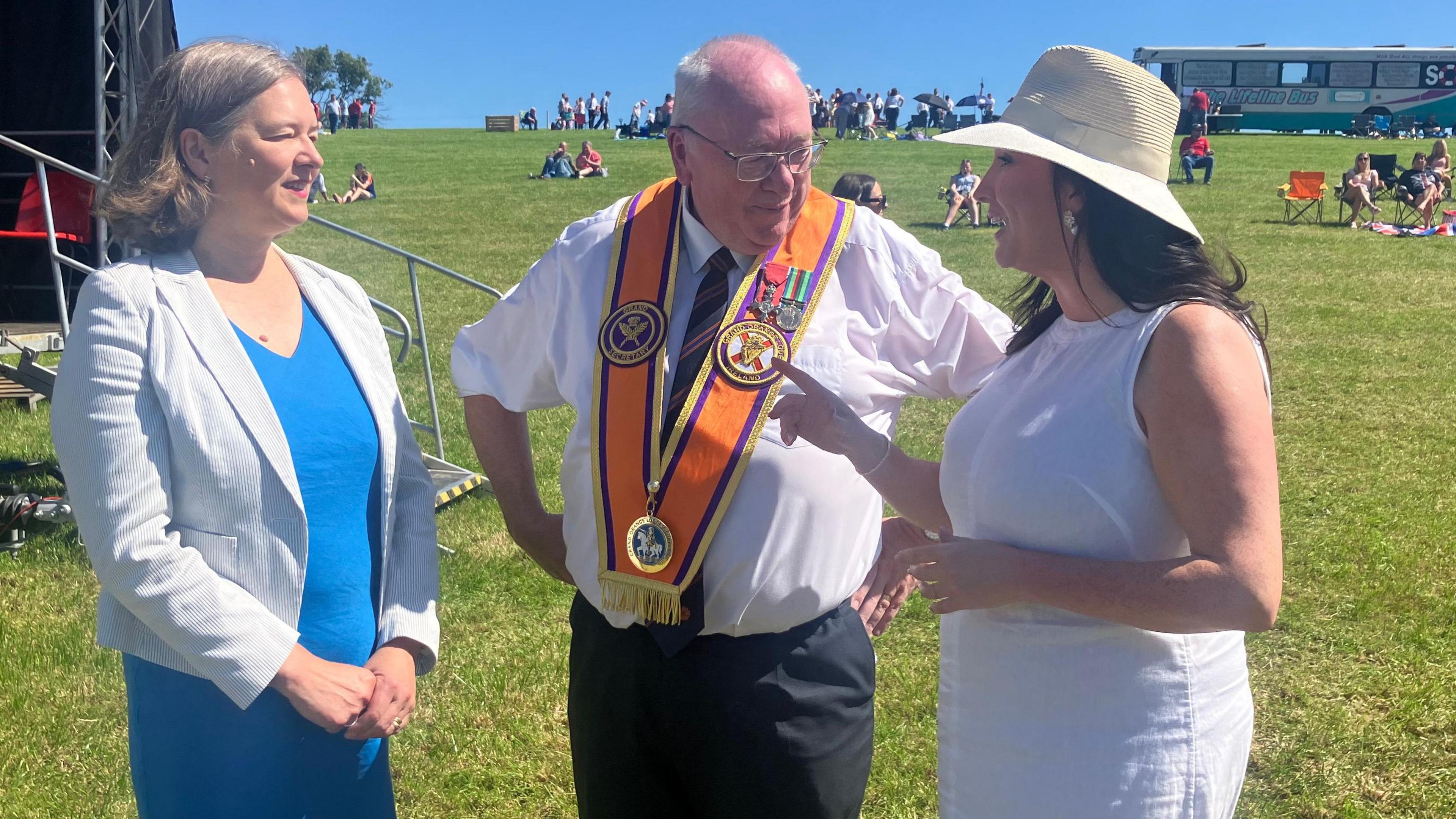 Fleur Anderson, Rev Mervyn Gibson and Emma Little-Pengelly standing on a field in the sun chatting to each other.