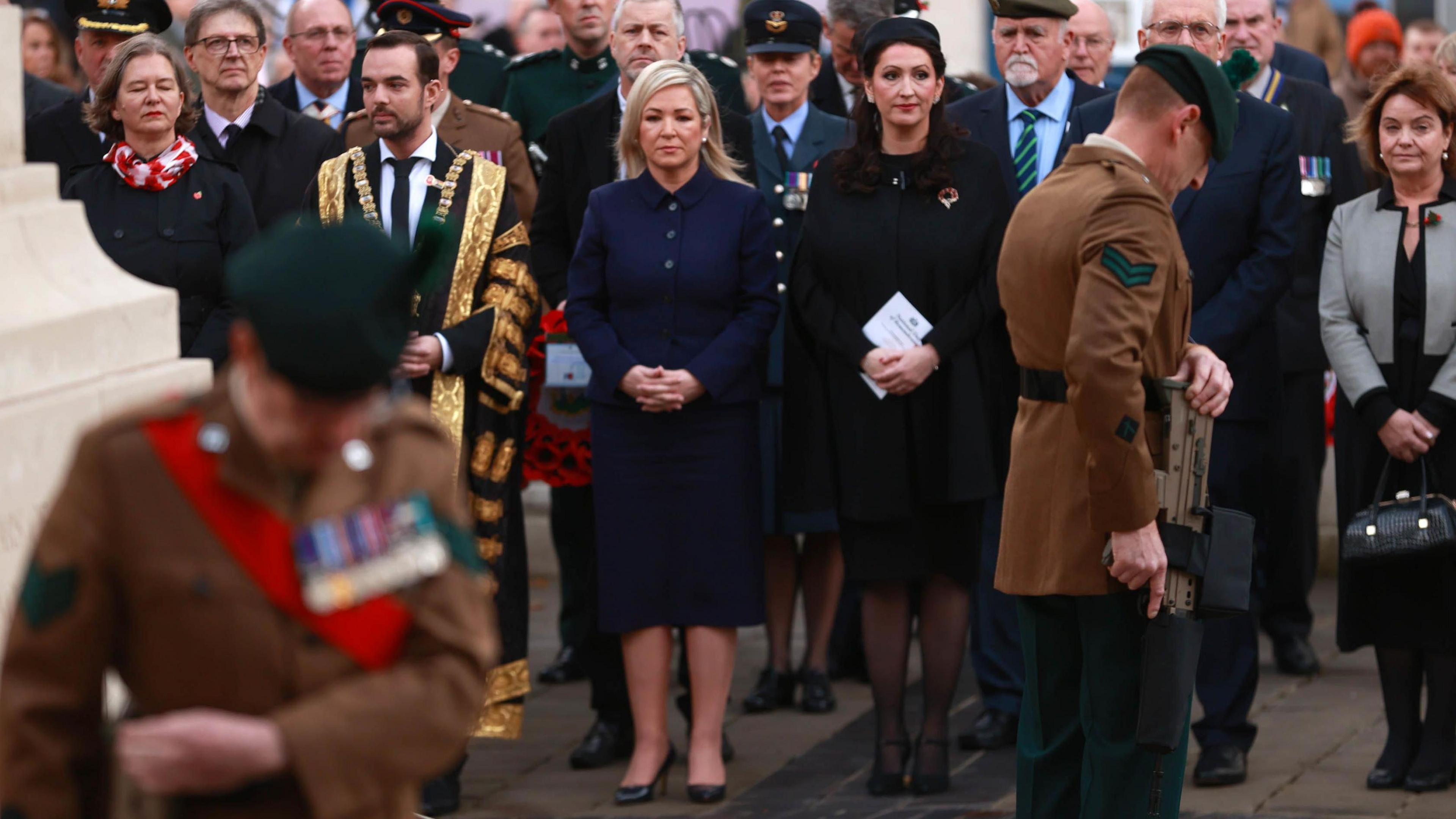 Michelle O'Neill and Emma Little-Pengelly standing together, both with their hands crosses in front of them. Michelle O'Neill is wearing blue and Emma Little-Pengelly is wearing black and has a poppy on her lapel. In the foreground a man in military dress who is out of focus.