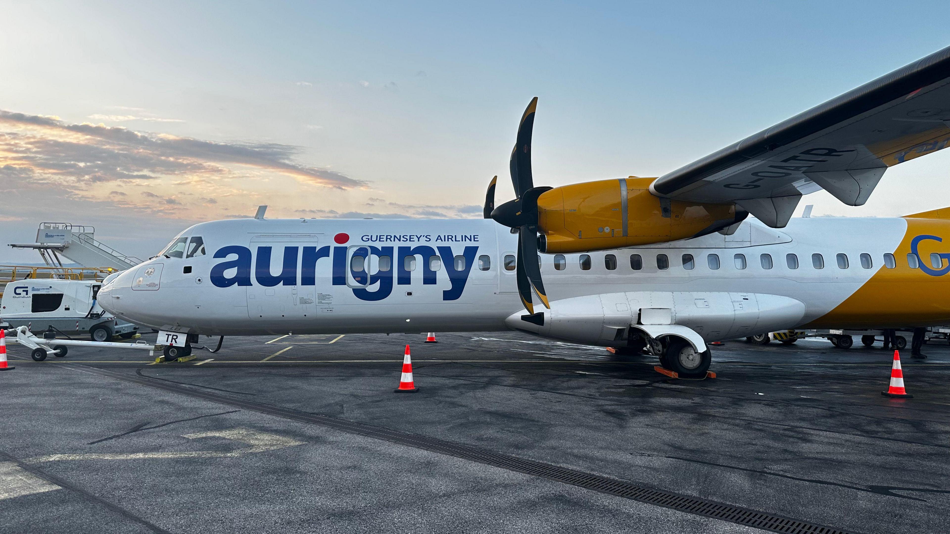 An Aurigny plane seen on the runway. It is white with yellow as its secondary colour and large black propellers. 