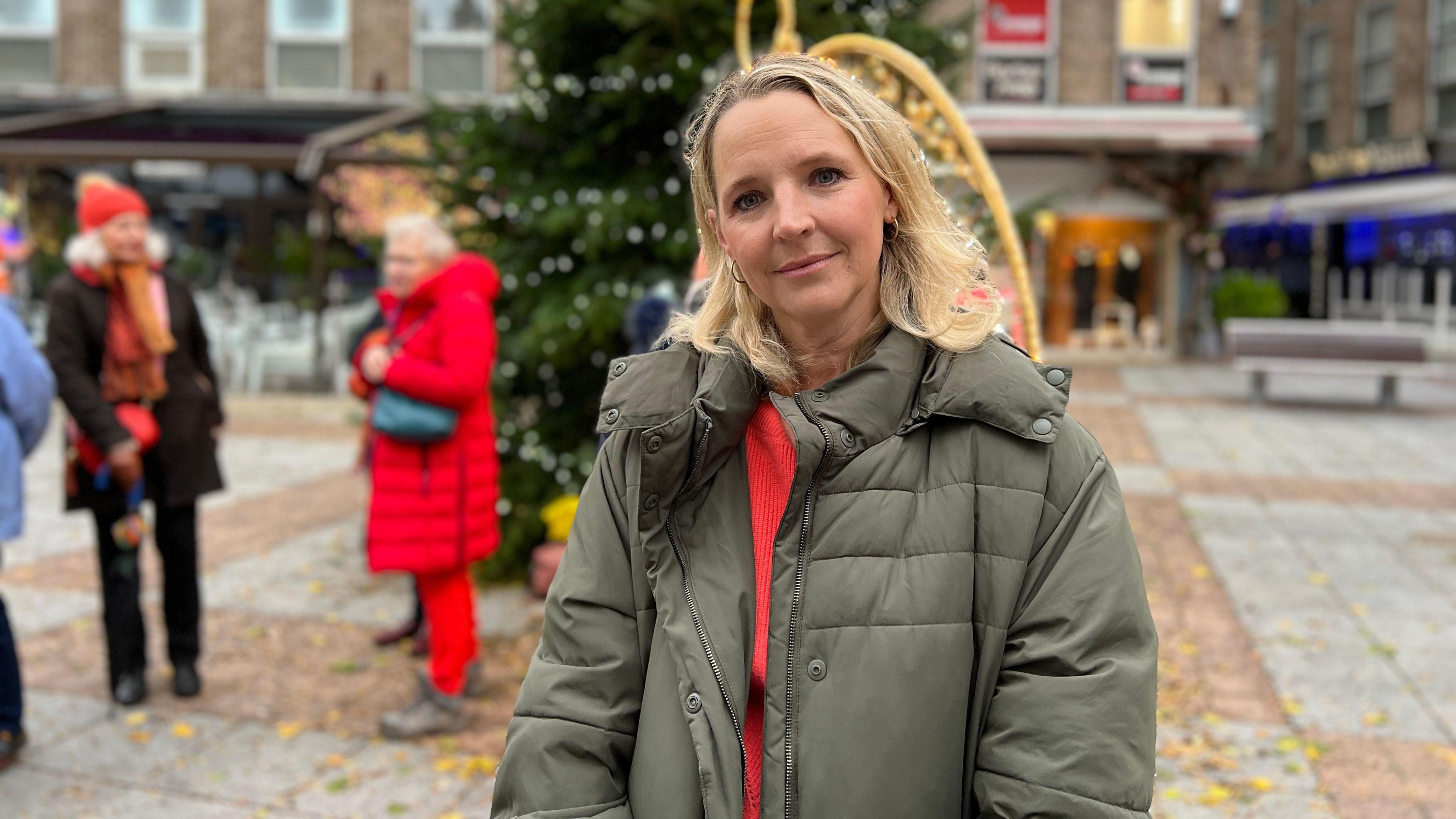 Kate looks at the camera as behind her a couple of women talk by a large Christmas tree in the square. Kate is wearing a light green coat with a red jumper underneath. She has short blonde hair which falls to her shoulders.