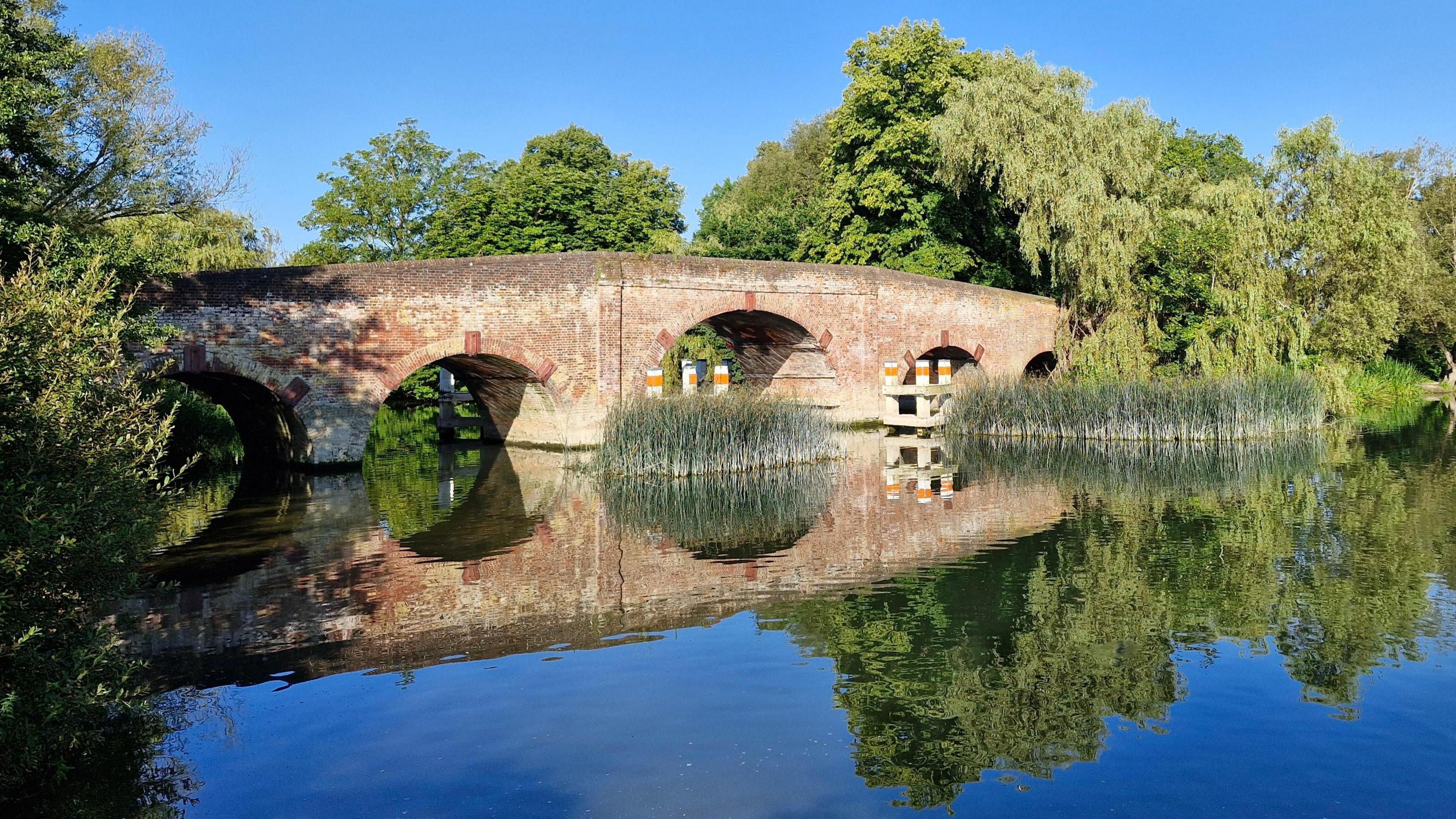 A old brick bridge over the river and it's reflection in the still water, Trees are on either side with reeds in front of two of the five arches