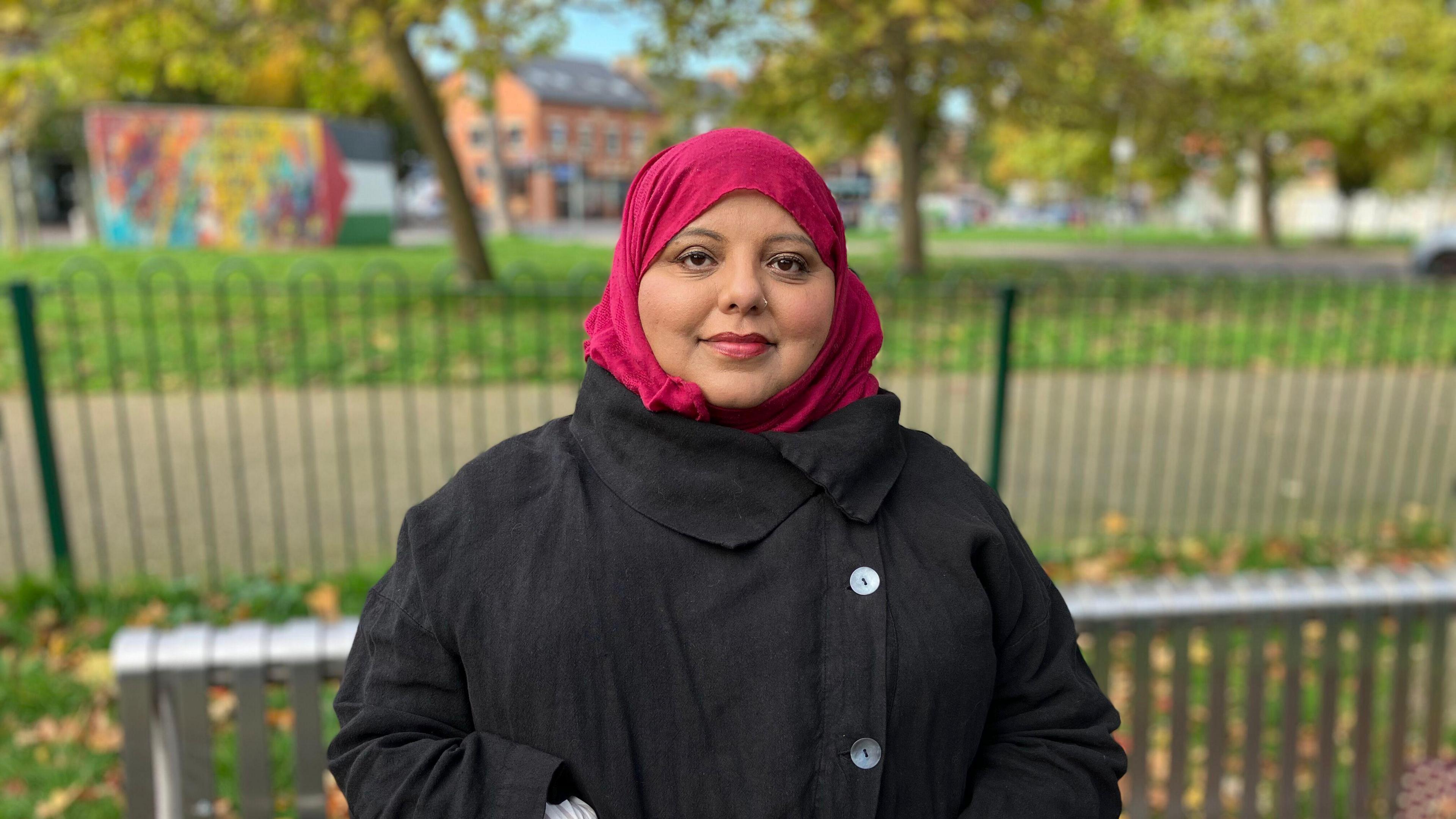 Shaista Aziz, who is one of the founders of the anti-racism campaign group The Three Hijabis, sits on a park bench in Oxford. She is looking directly at the camera and wearing a black buttoned up coat and a pink head scarf. 