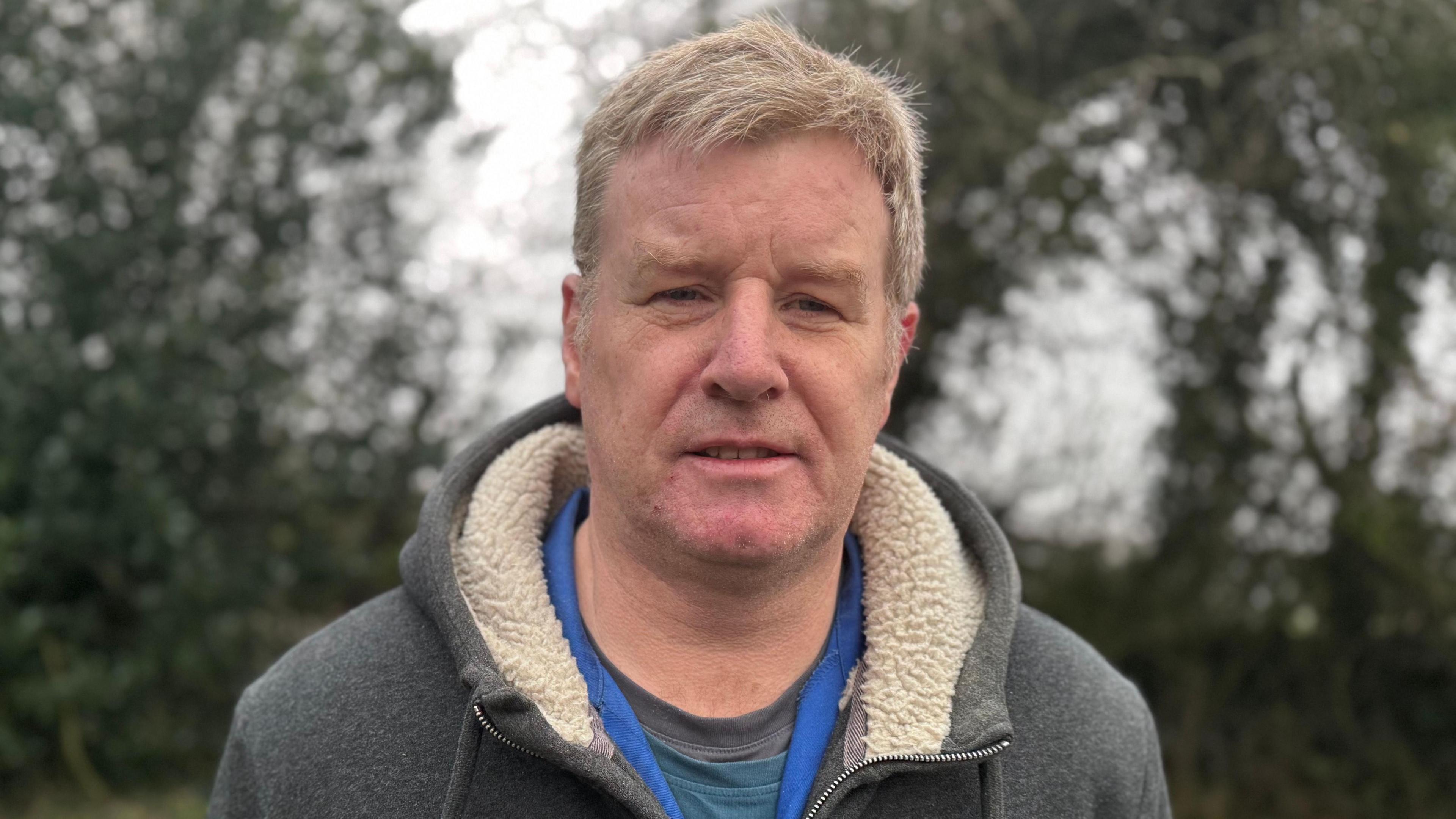 Mark Robinson - a man with short light hair looks at the camera. He is stood in front of some trees which are blurred in the background. He is wearing a grey cardigan with a woolly hood, a grey and blue tshirt and a darker blue collar.