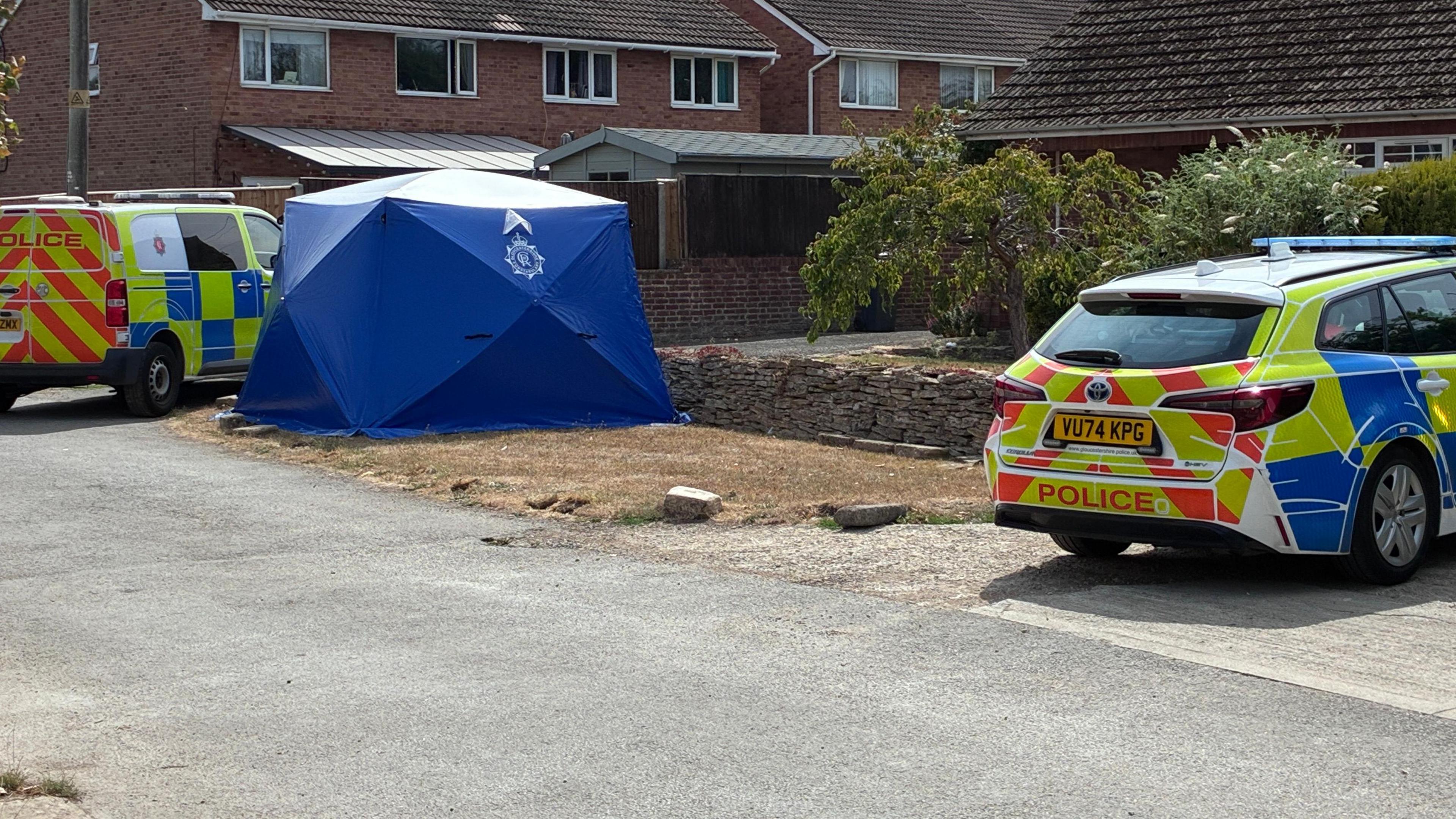 A blue forensics tent is on a grass verge by the side of a house with two police cars parked either side.