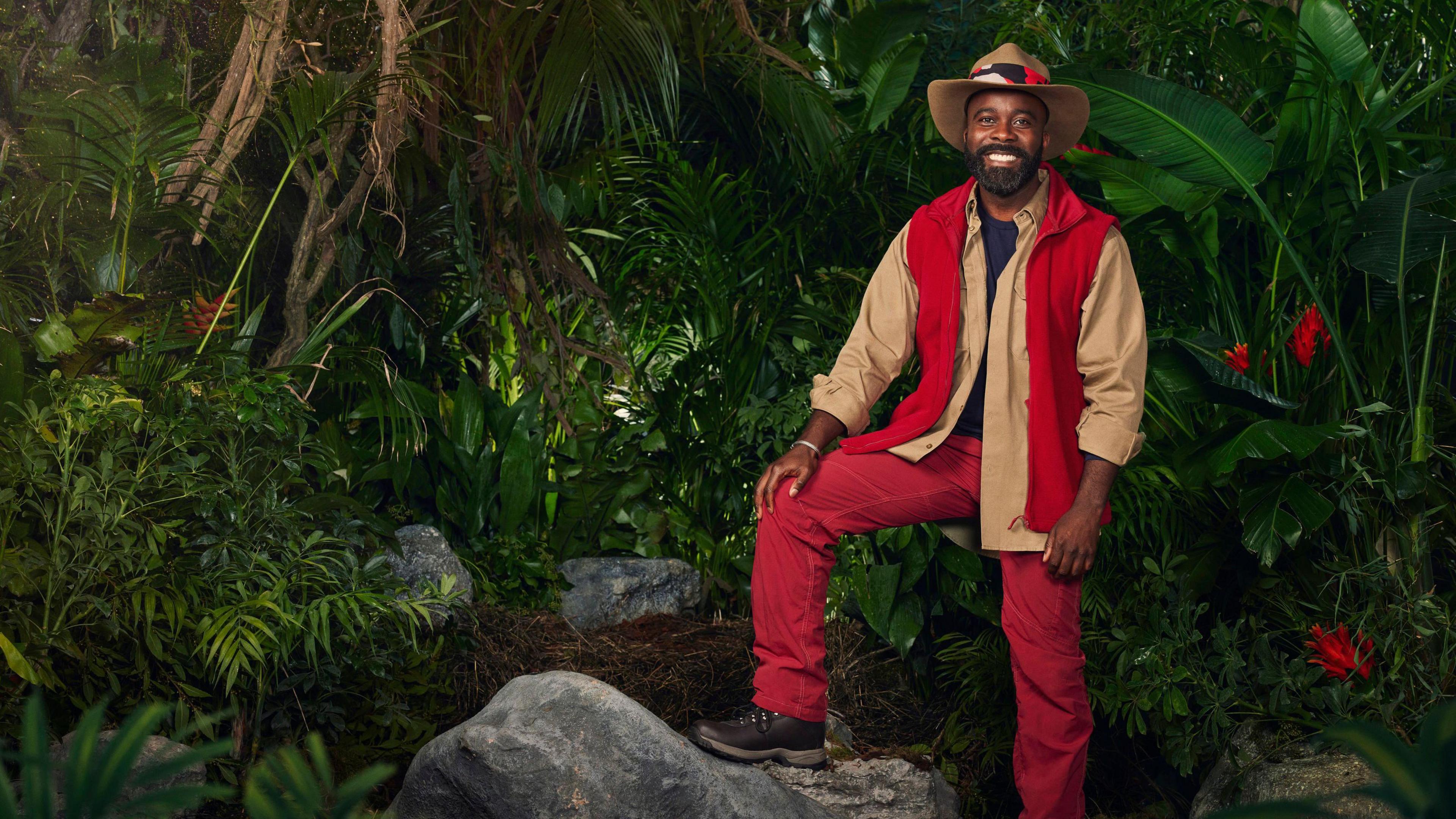 Melvin Odoom wearing his jungle outfit, standing with his foot on a rock in a jungle scene 