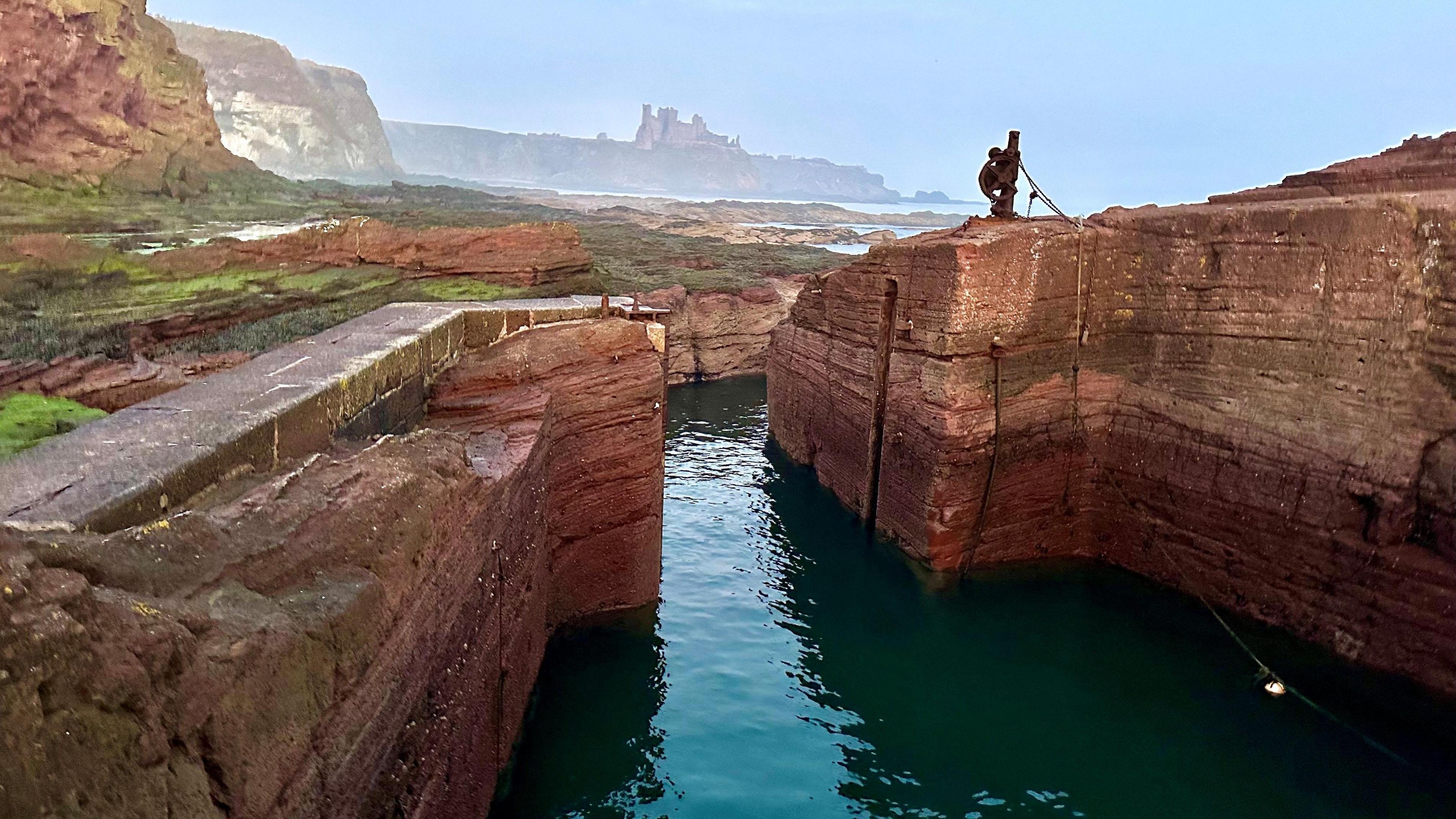 A general view of Seacliff Harbour in East Lothian. The harbour has red and orange sandstone on either side. There is blue water in the middle. Patches of green moss are on the left side of the image. The view of Tantallon Castle, high up in the background, is shrouded by a light mist.