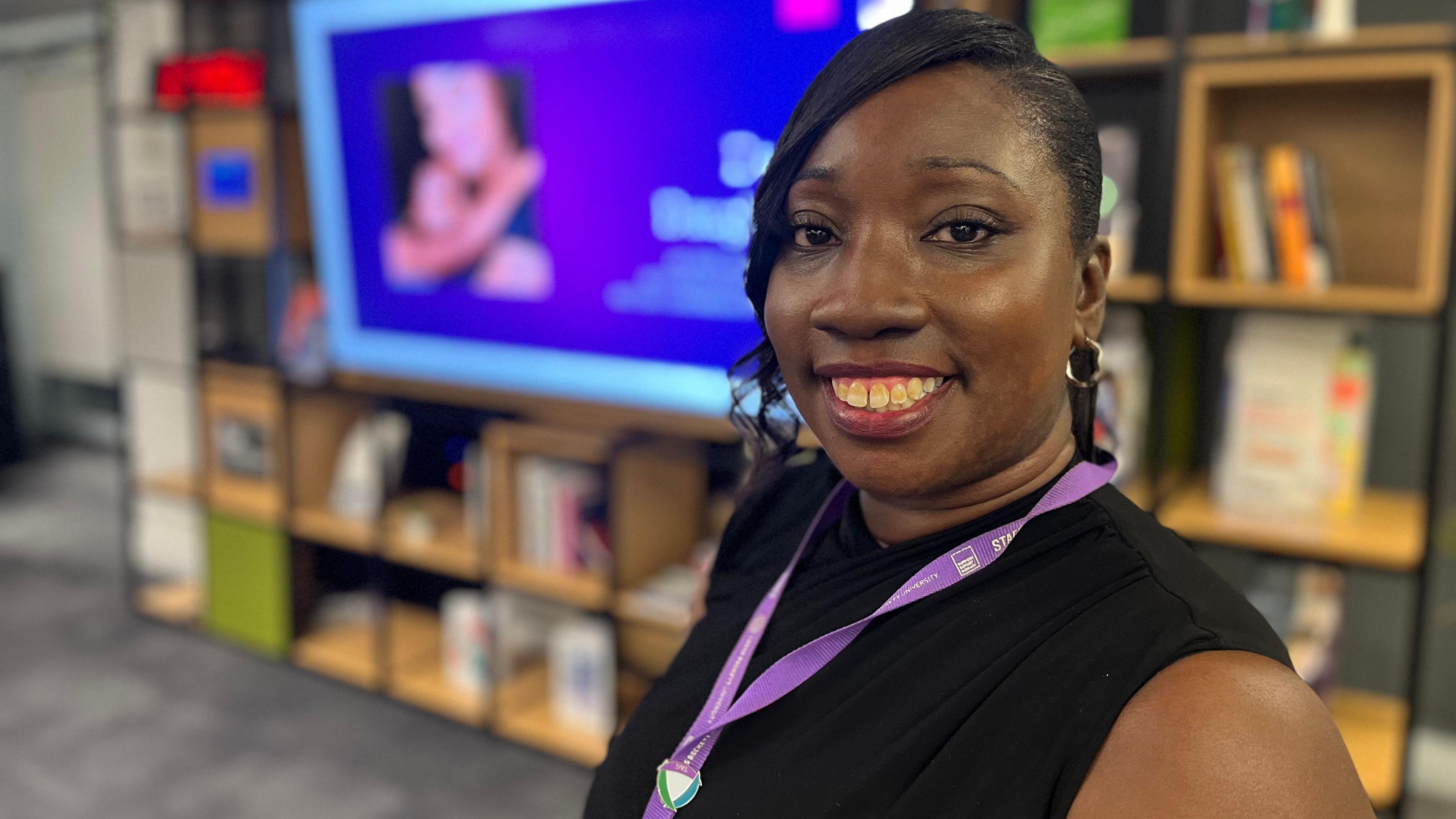 Shirleeccia Ward from Leeds Beckett University stands in front of a screen in a library, wearing a black top and a purple lanyard 