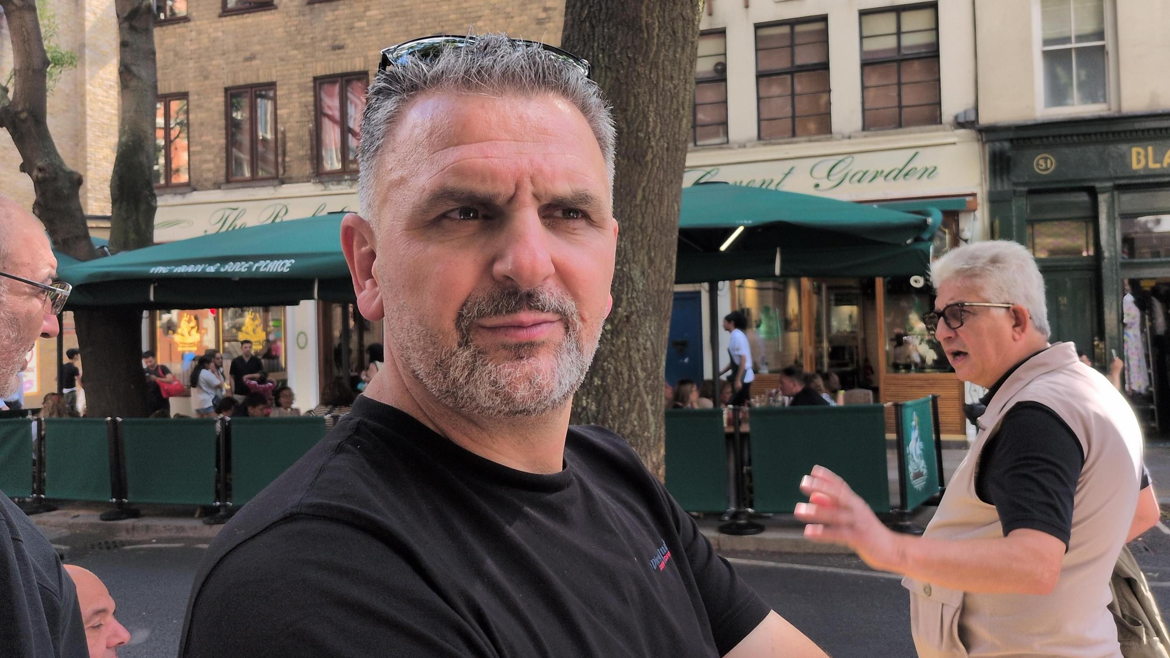 A man in a black shirt with sunglasses on his head stands in the streets.
