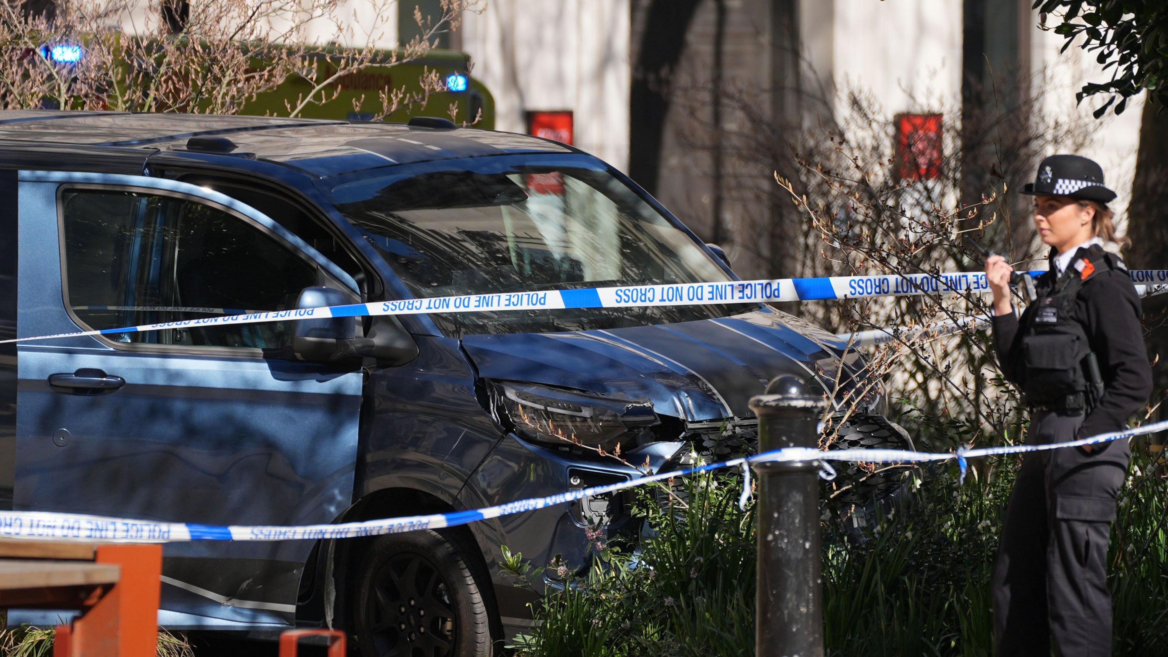 A blue van with a black stripe running across its roof and bonnet is parked behind blue and white police tape in The Strand. A uniformed officer can be seen speaking into a radio