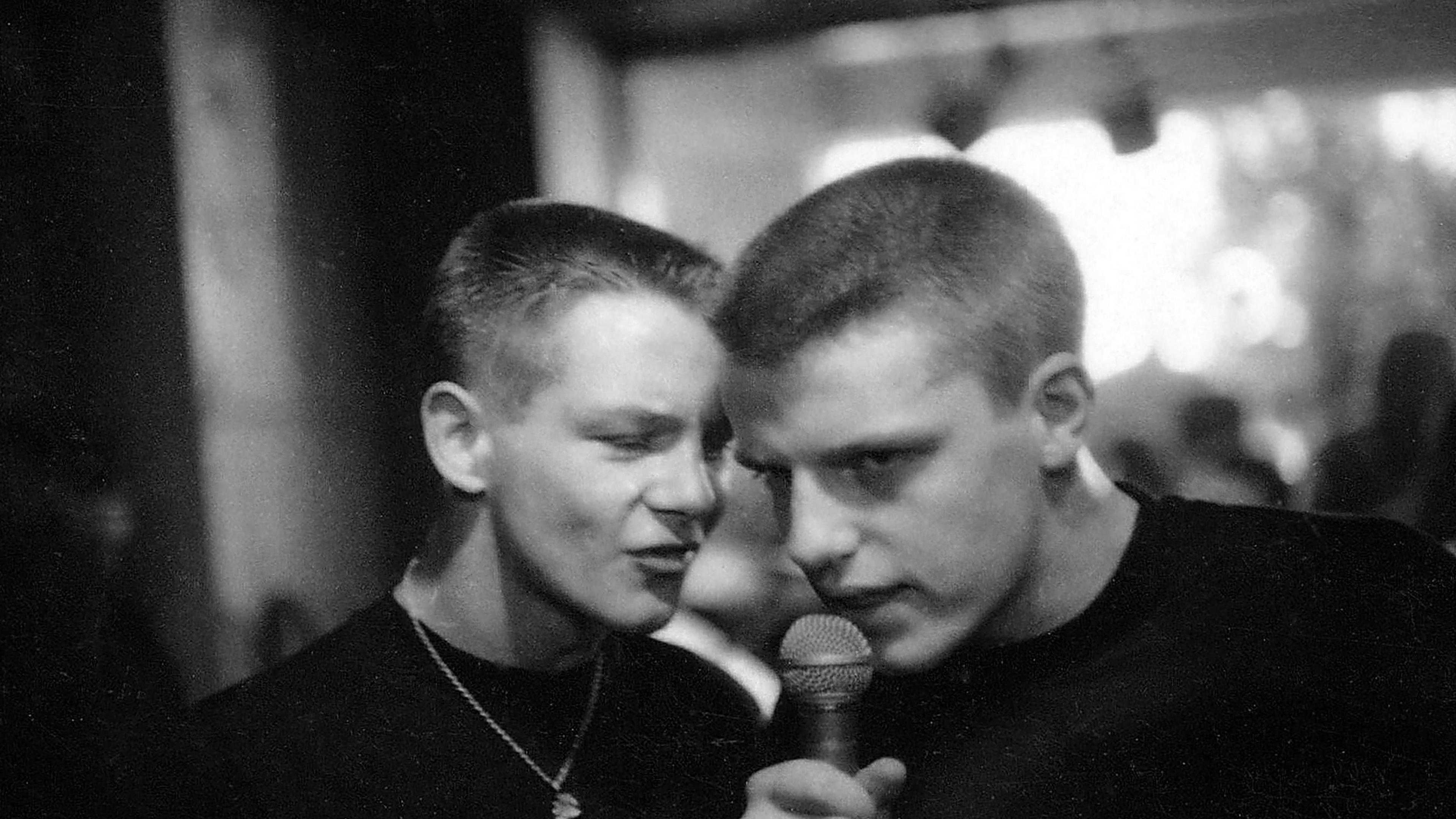 Two members of the band Madness pictured in black and white in close proximity to each other. One is talking or singing into a microphone.