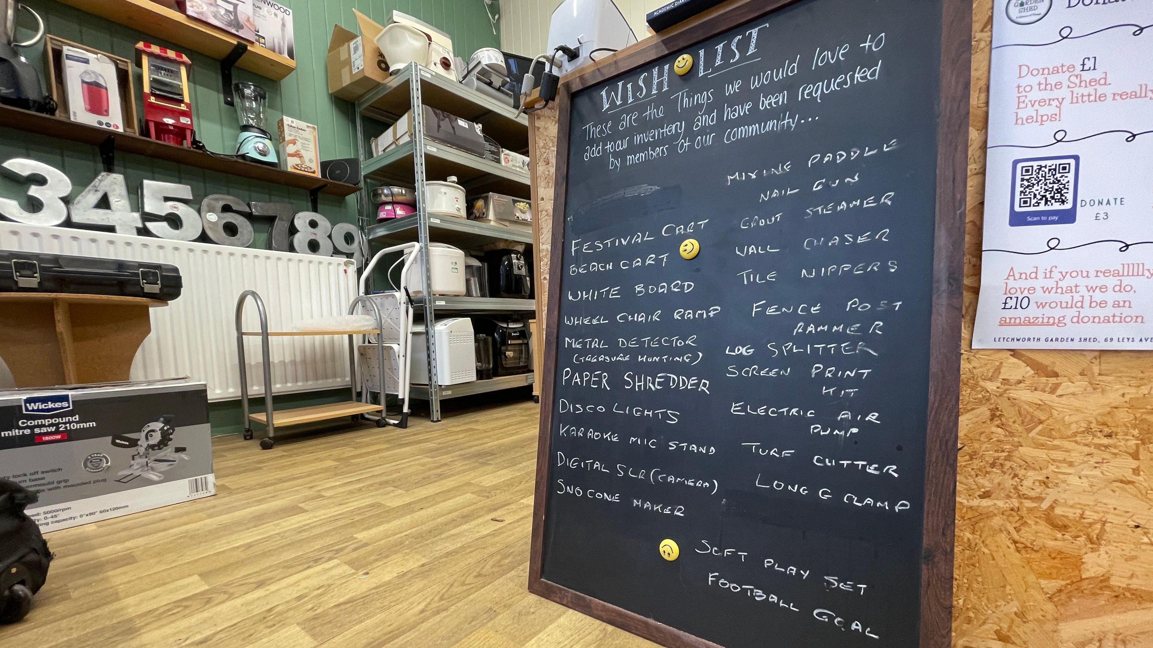 A blackboard is propped against the front till of the Letchworth Community Shed. In white chalk it reads: Wish List. These are the things we would love to add to our inventory and have been requested by members of our community: festival cart, beach cart, white board, metal detector for treasure hunting, paper shredder, disco lights, karaoke mic stand, digital SLR camera, snowcone maker, mixing paddle, nail gun, grout steamer, wall chaser, tile nippers, fence post rammer, log splitter, screen print kit, electric air pump, turf cutte, long G-clamp, soft play set and football goal.