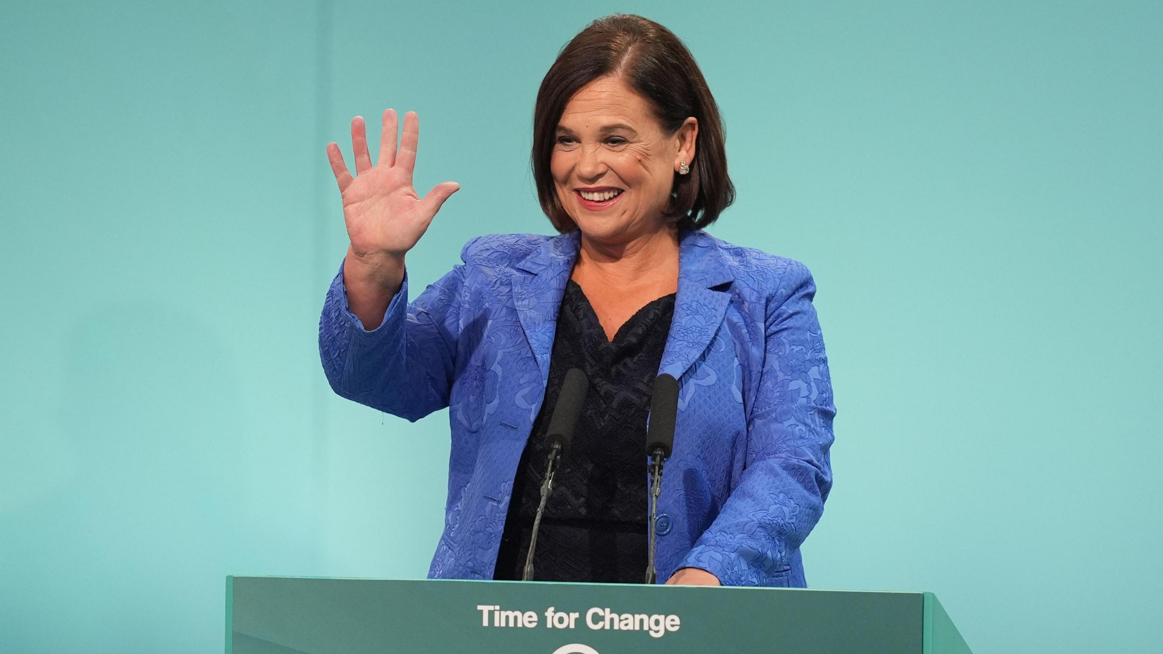 Sinn Fein's President Mary Lou McDonald standing at a light green podium giving her address at Sinn Fein Ard Fheis. She is wearing a blue patterned suit jacket and a black top. 