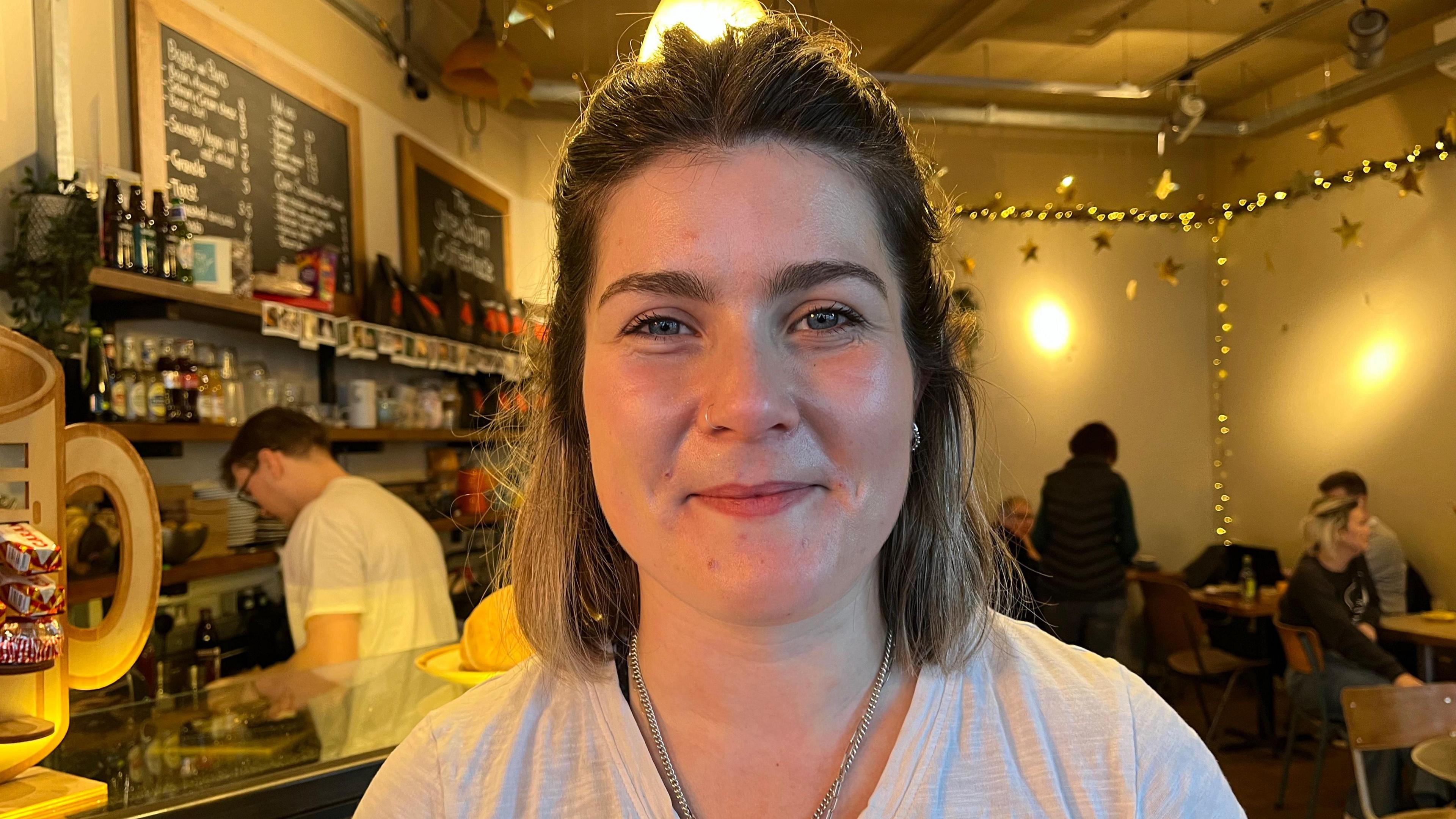 Livi looking into the camera and slightly smiling. Behind her you can see the Shrewsbury Coffeehouse, it's a busy cafe with people sat at tables and chatting. Over the other shoulder you can see the till and coffee machines. There's fairy lights and lamps which give the room a warm glow.