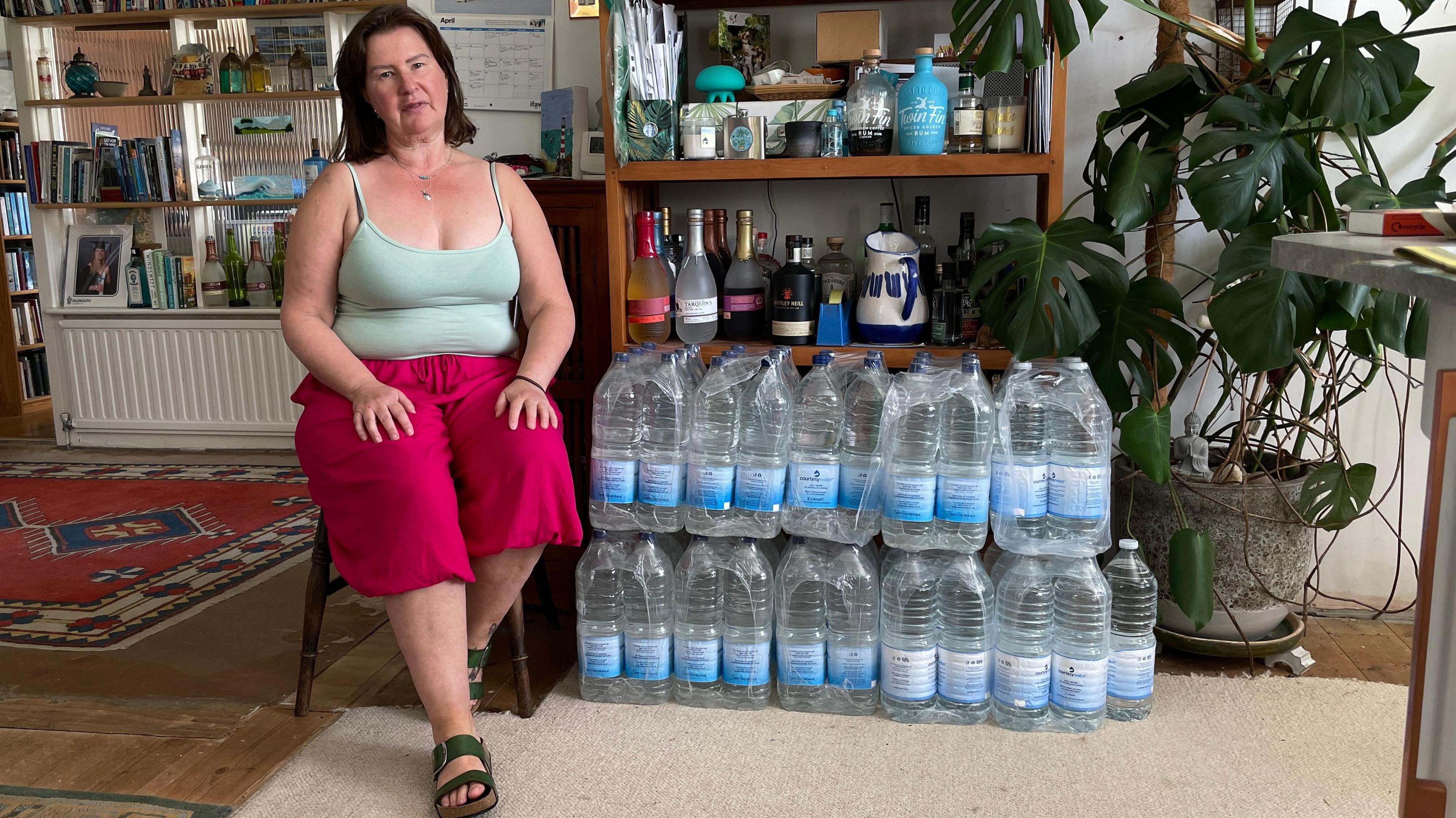 A woman is sitting on a wooden chair in her kitchen. She is wearing a light green tank top and bright pink shorts. Next to her are rows of water bottles stacked on top of each other. In the background there are bookshelves with some books, plants, and other household items. There are also several bottles and containers on the shelves as well as a large green house plant.
