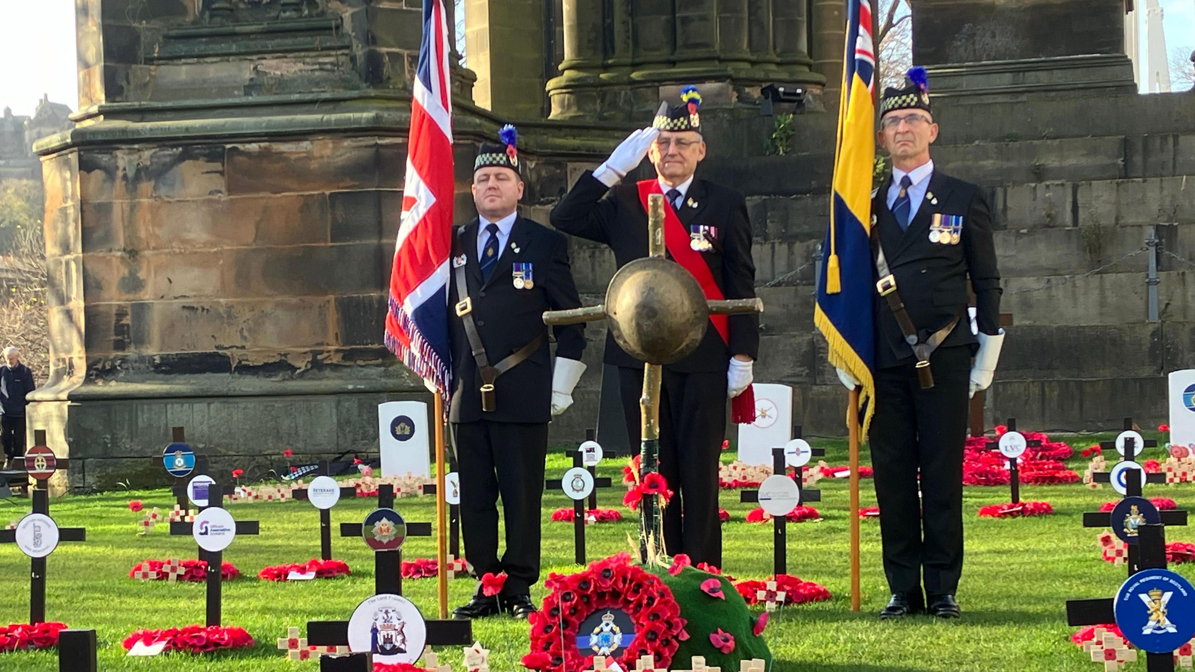 Three uniformed men stand in a Remembrance garden in Edinburgh Princes Street flanked by two flags, one of which is a Union Flag. They are all saluting and there are small crosses on the ground all around them. 