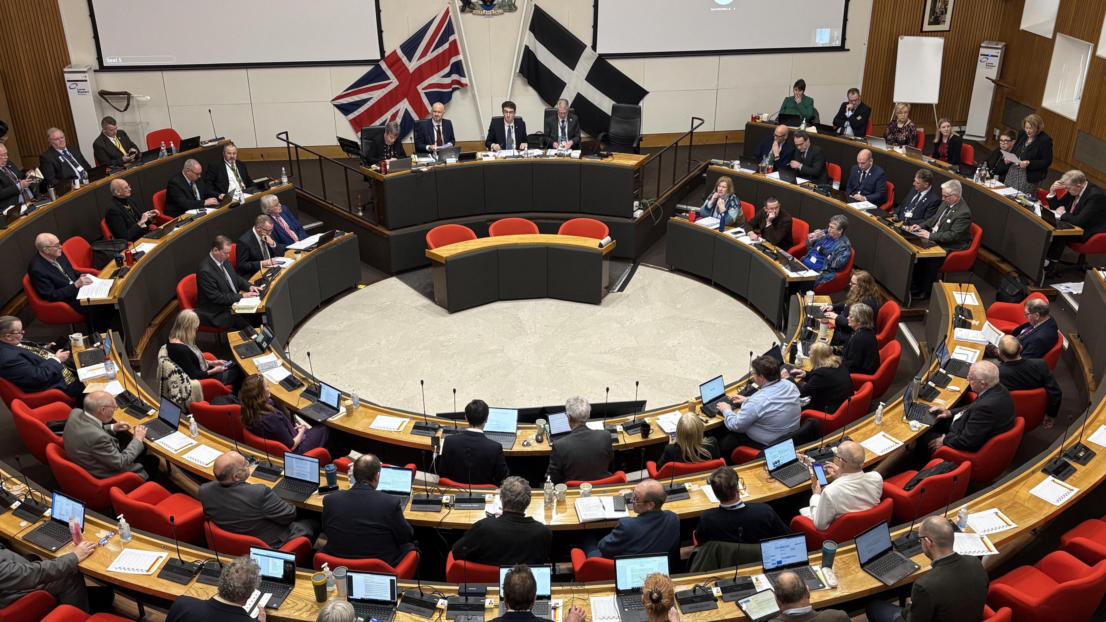 Councillors sitting in the chamber at Cornwall Council. The large, circular, wood-panelled chamber is filled with numerous individuals seated in red chairs. Many attendees have laptops, documents and microphones in front of them. At the front of the chamber is a raised platform where a panel of three individuals is seated behind a central desk. Behind them are two flags: the UK's Union Flag and the black flag with a white cross of Cornwall. 