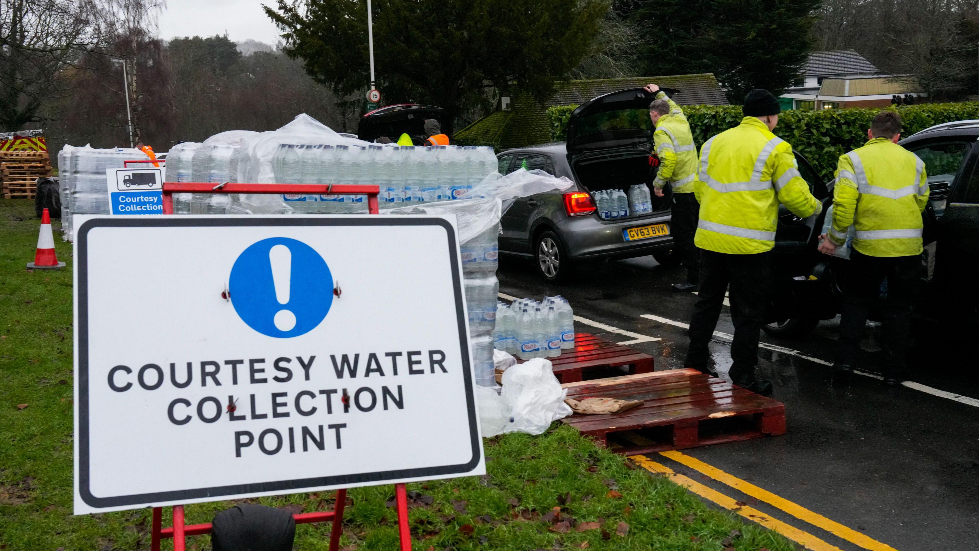 A sign saying "courtesy water collection point" is in the foreground. There are piles of bottled water behind it and cars queueing on the right of the image. Bottles of water are being put in the boot of cars by people in yellow high vis jackets
