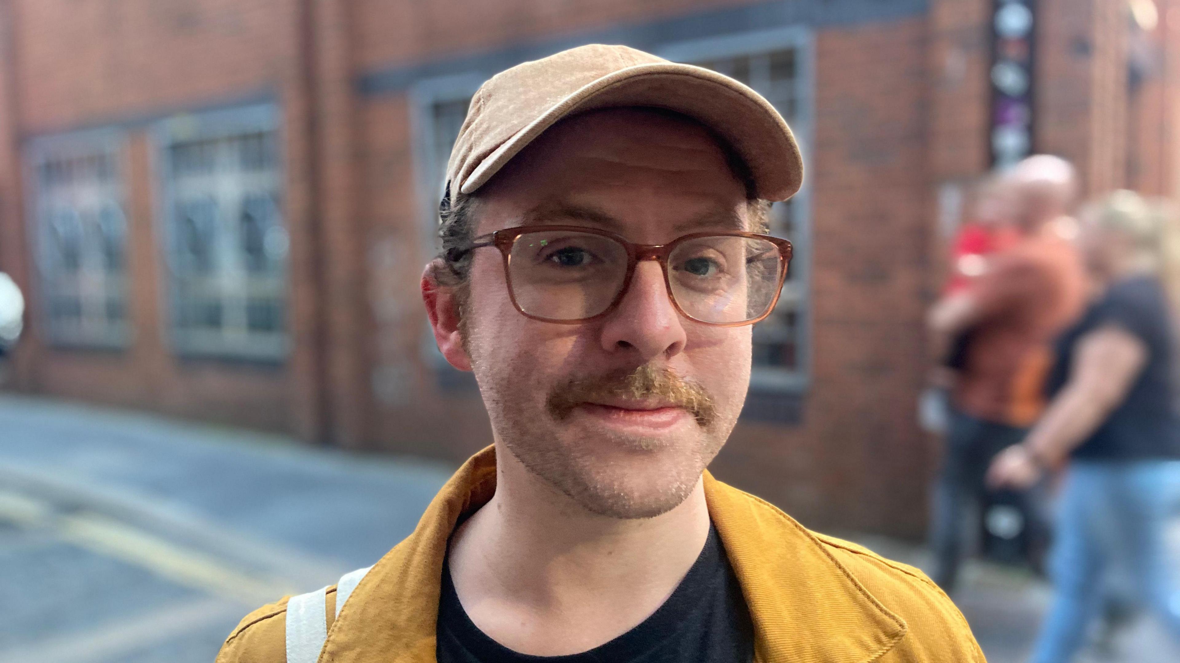 Matt, a man with short hair and a fair to ginger moustache, standing in a street in Belfast. He is wearing a peaked cap, a black t-shirt and a yellow jacket. 