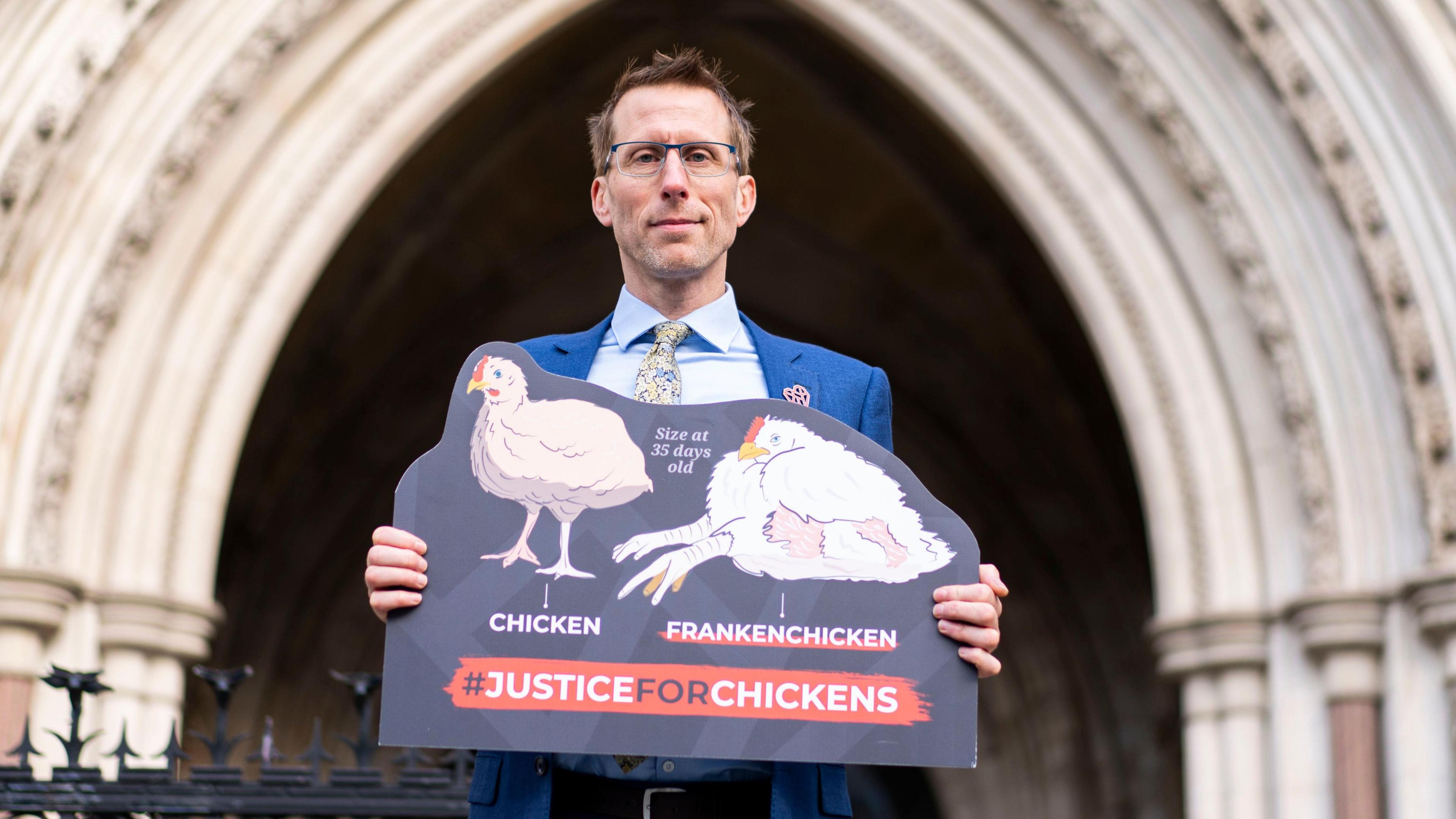 Mr Gifford holds a placard outside the gates of the court.  The placard reads Justice for Chickens - and shows two illustrations of a normal chicken against a frankenchicken at 35 days old - the larger frankenchicken is unable to stand.