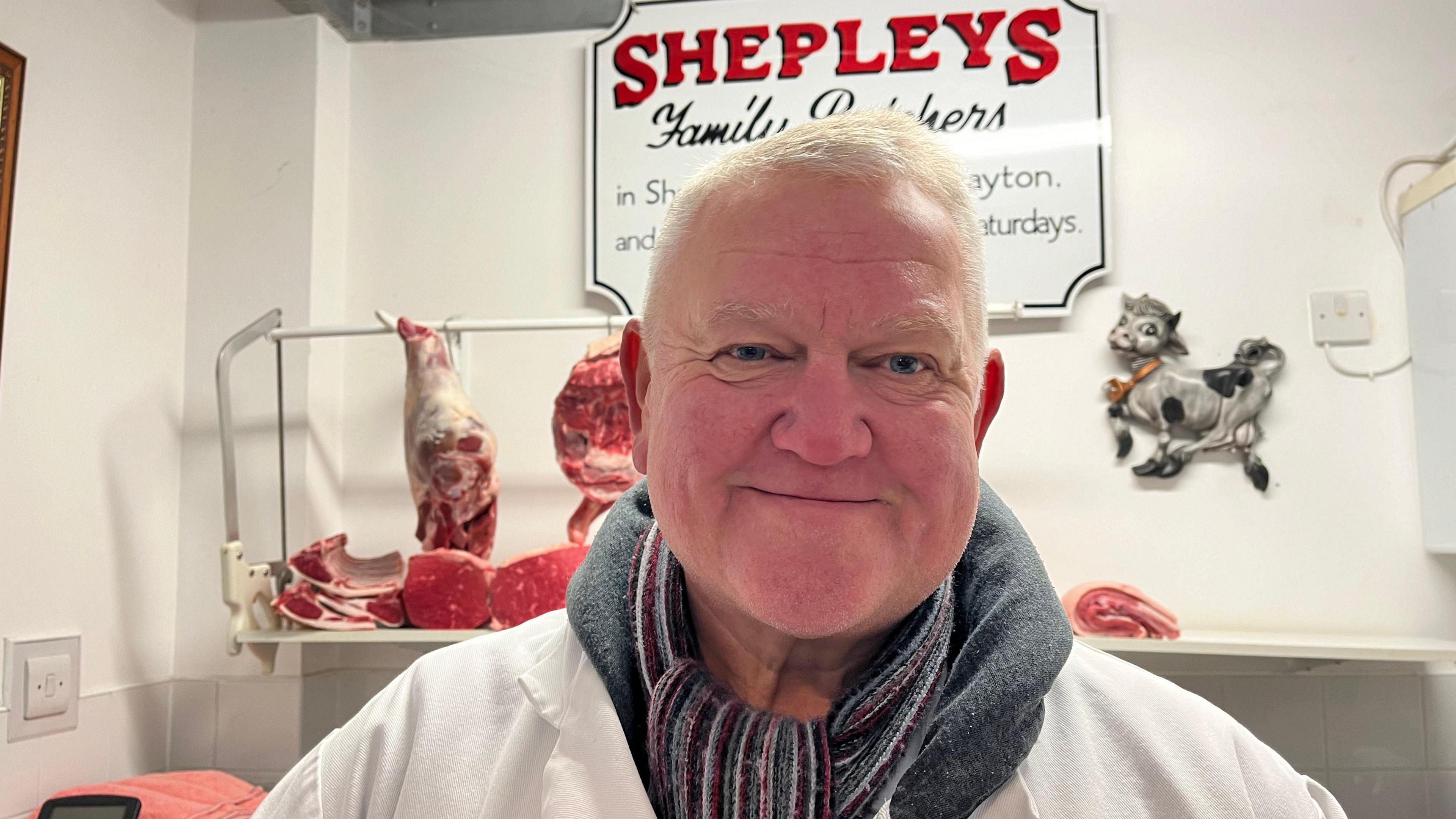 Richard is wearing a white coat over the top of a winter jacket and scarf. Behind him there's meat hanging up ready to be sliced up, and a sign that reads 'Shepley's Family Butchers.'