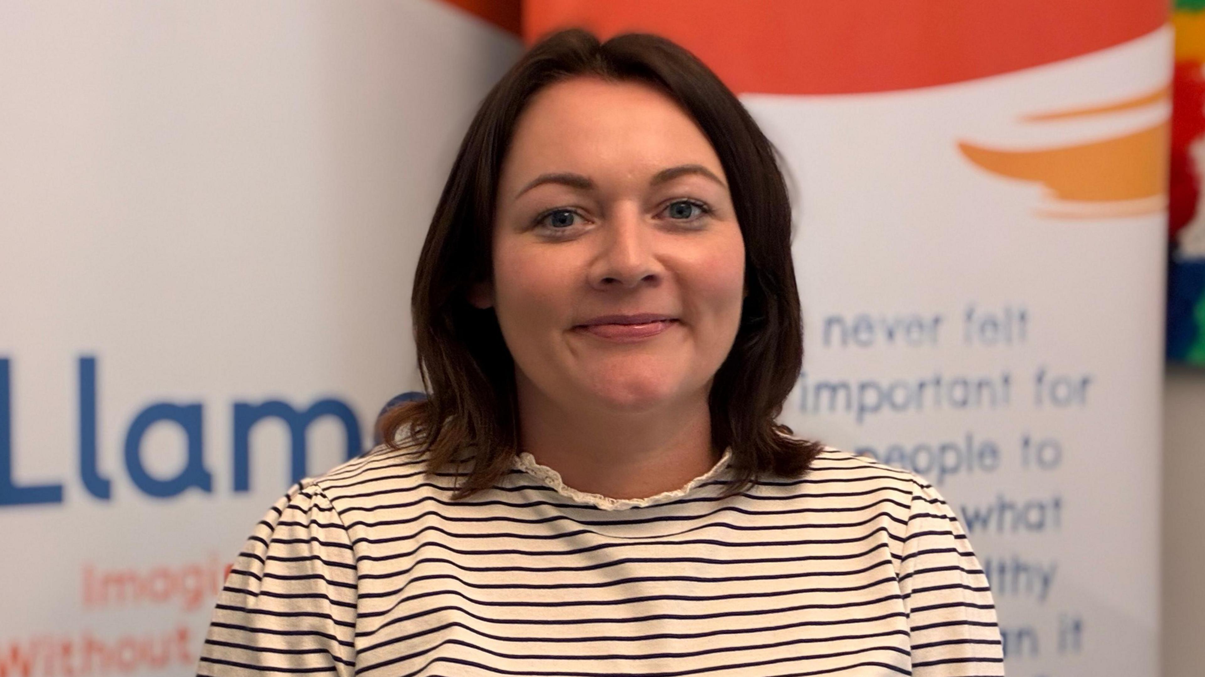 Yvonne photographed indoors in front of promotional display banners, wearing a striped long-sleeved top.
Yvonne has shoulder length brown hair and is smiling at the camera.