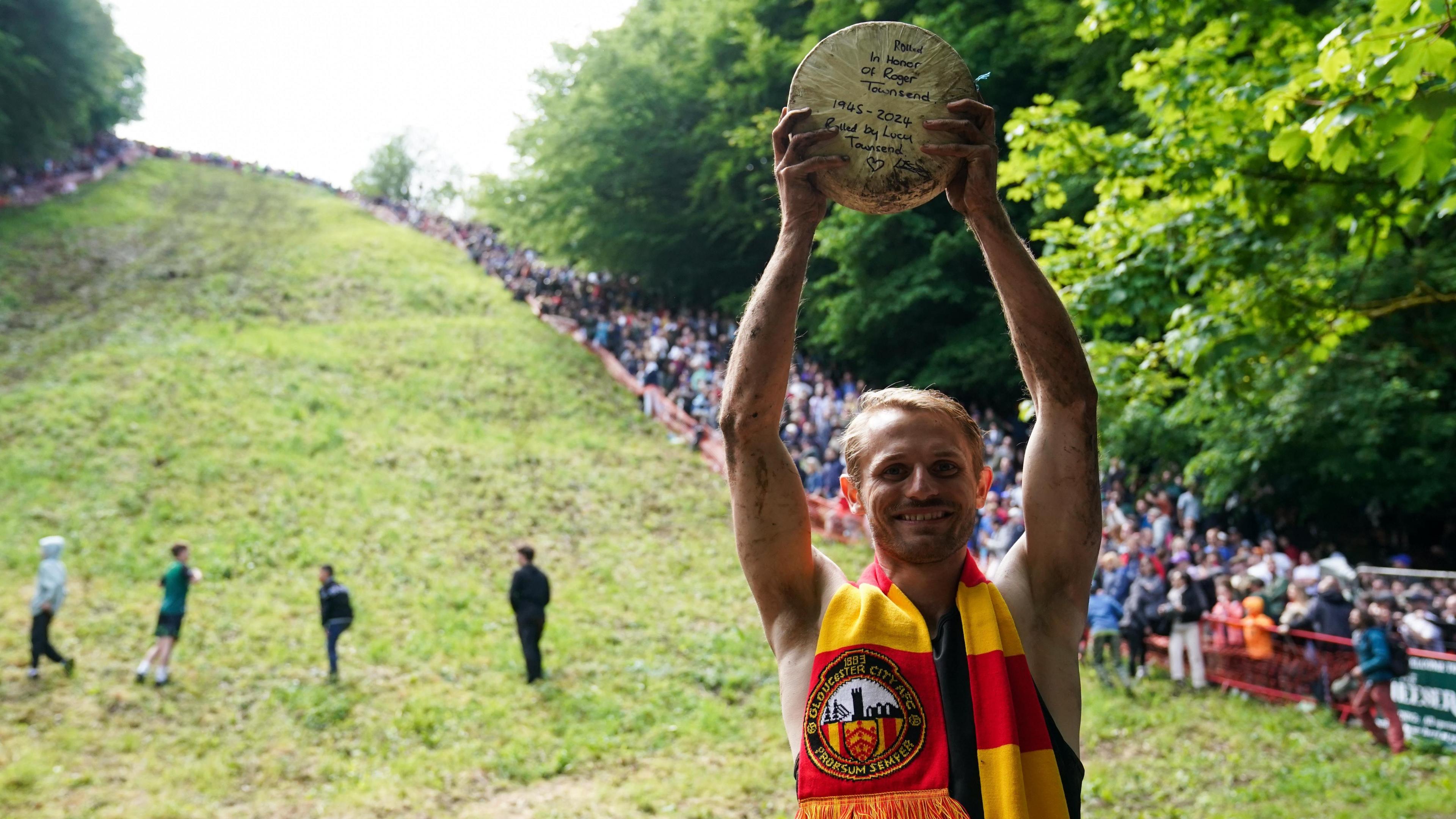 Dylan Twiss holding up a cheese wheel at the bottom of the hill, wearing a Gloucester City FC scarf