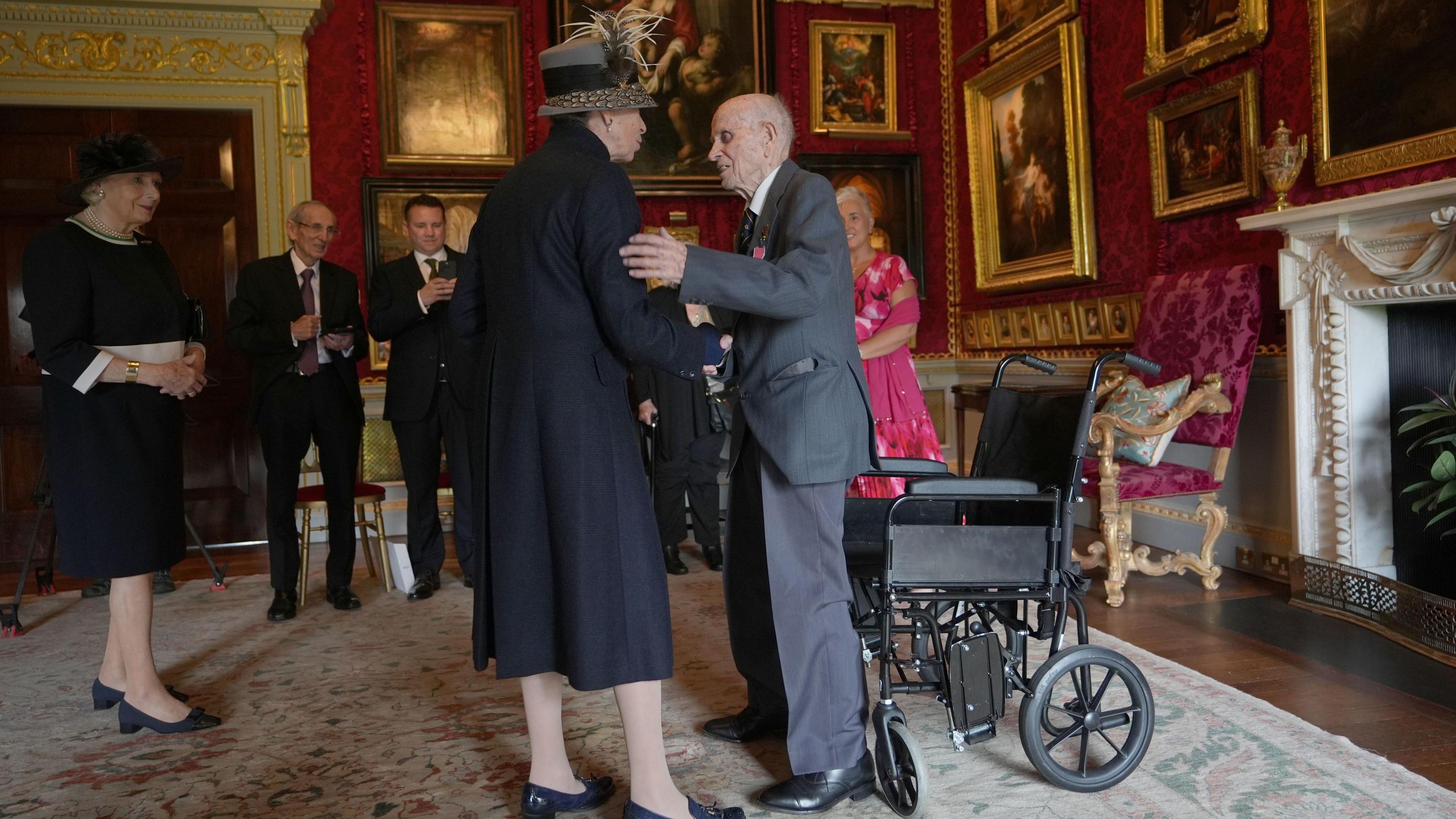 The Princess Royal wearing a black coat and grey hat inside a room in Hillsborough Castle. She has a bag over her shoulder. She is speaking to Norman Irwin, who is in a grey suit. He is standing, with his wheelchair behind him. He has a medal in his pocket. There are gold-framed photos on the walls and a large fireplace. Four people are standing round the edge of the room, smiling.
