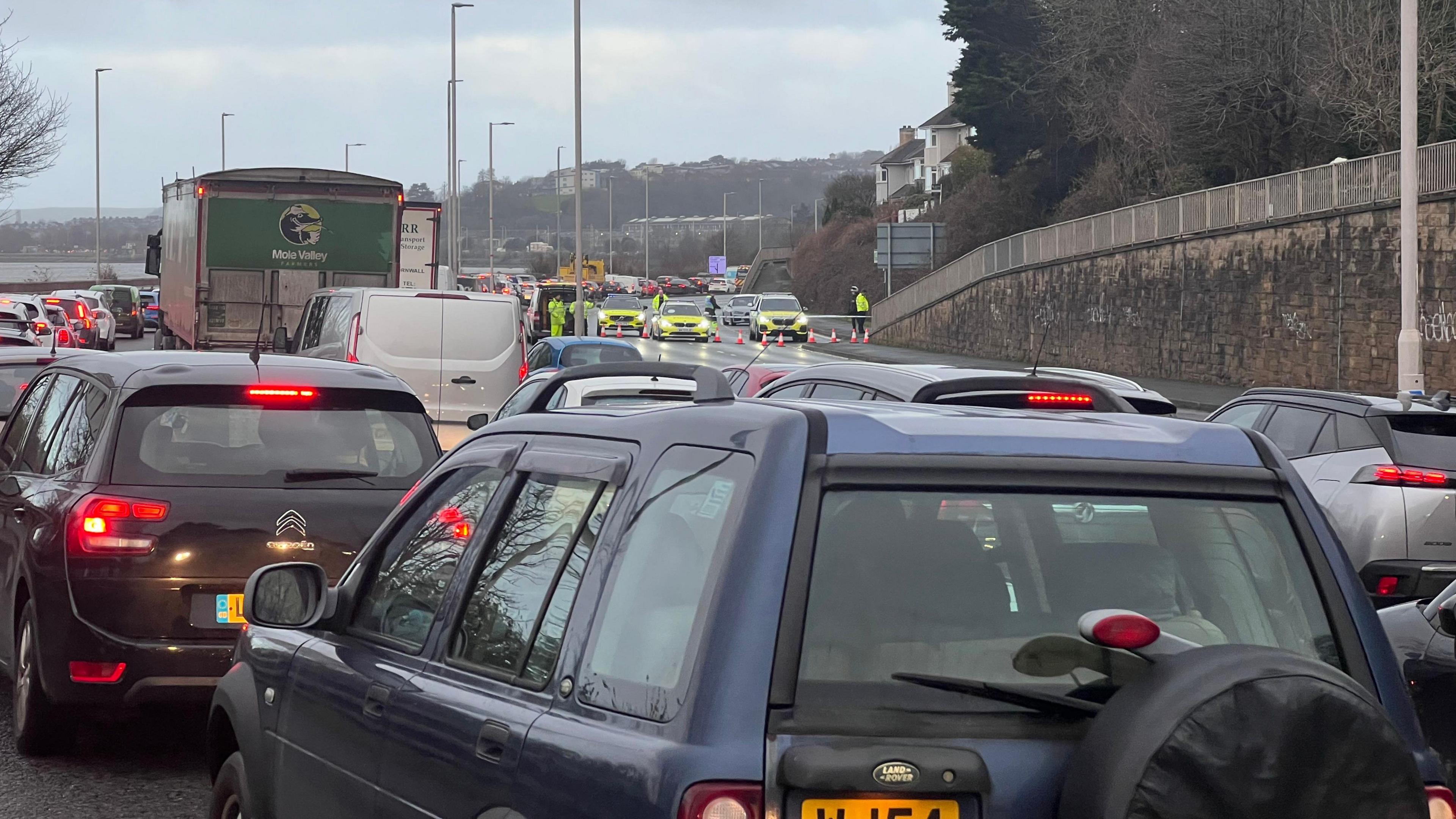 Traffic queuing along a road in Plymouth. Traffic on the left hand side of the road is queuing and there is a police closure on the right hand side of the road. 