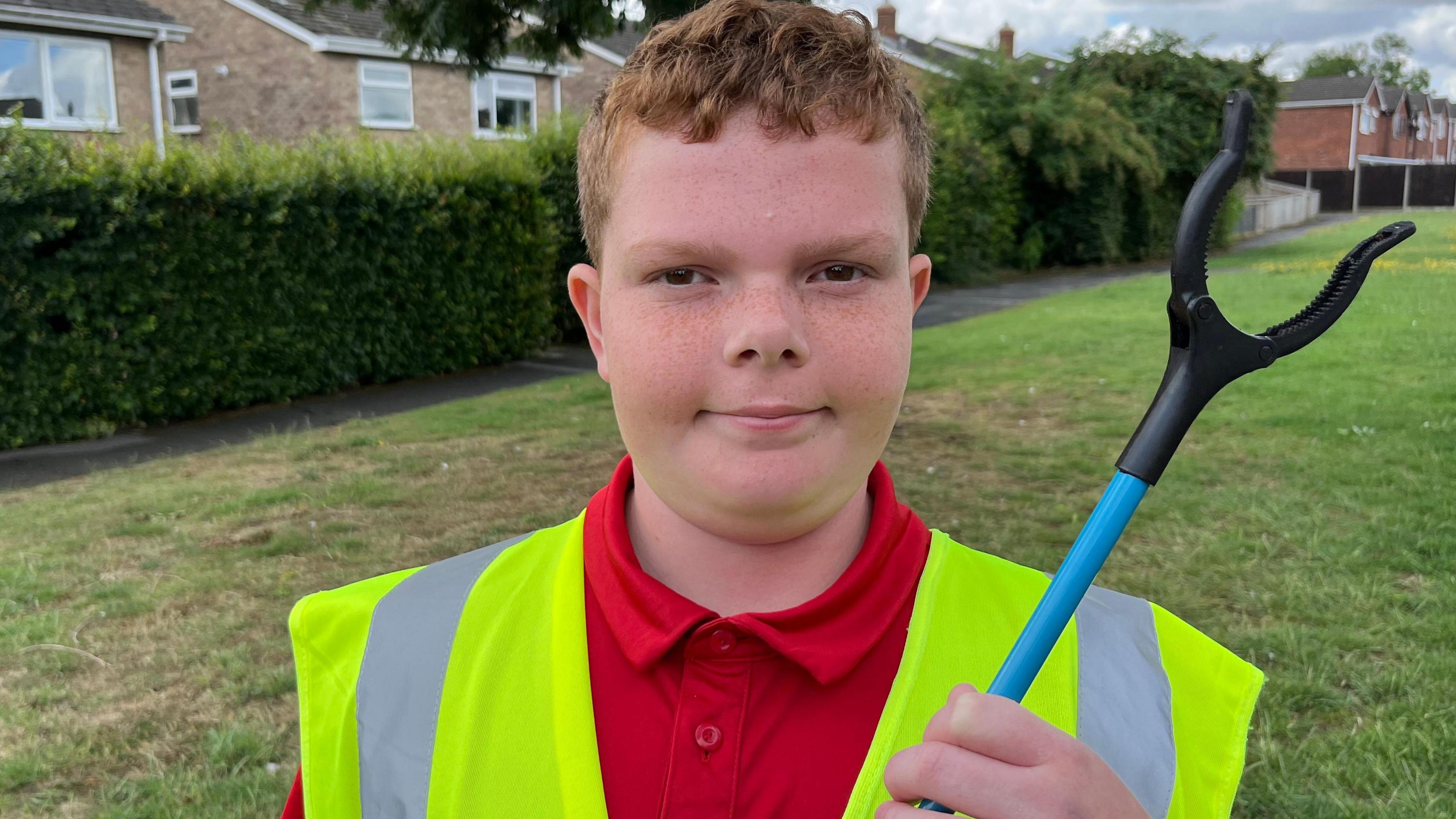 A head and shoulders shot of a boy with short gingery hair, wearing a hi-vis yellow waistcoat over a red polo-shirt. He is holding a blue-handled litter picker up to his shoulder. Behind him is grass, a hedge and some just glimpsed houses.