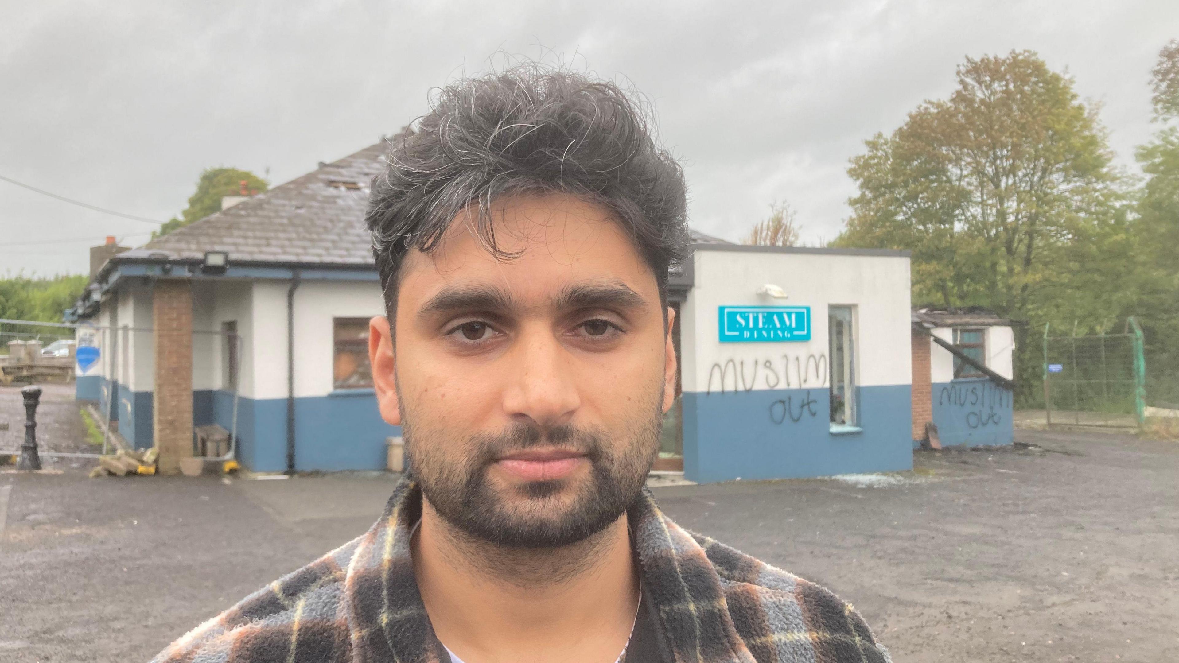 Abjan Acharya is standing in front of his arson-hit restaurant, with graffiti saying Muslim Out visible on a wall. He has dark hair and dark stubble and is wearing a checked top