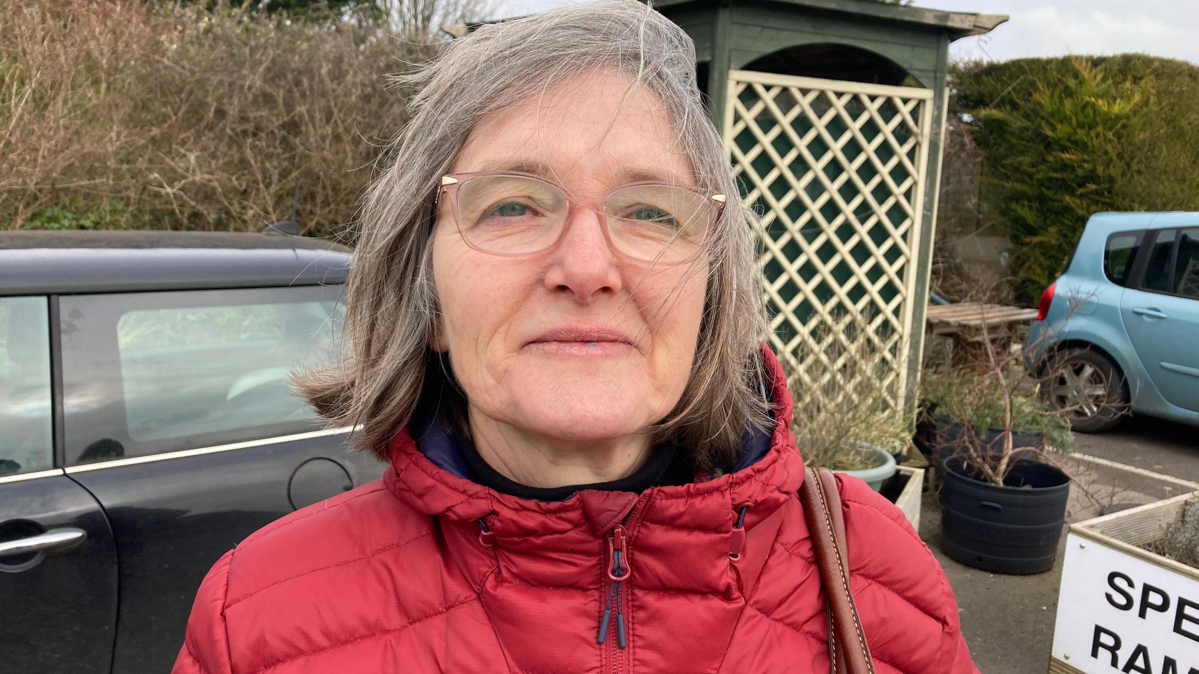 Rosie Dawson is standing in front of some cars in a car park. She is wearing a red jacket and glasses.