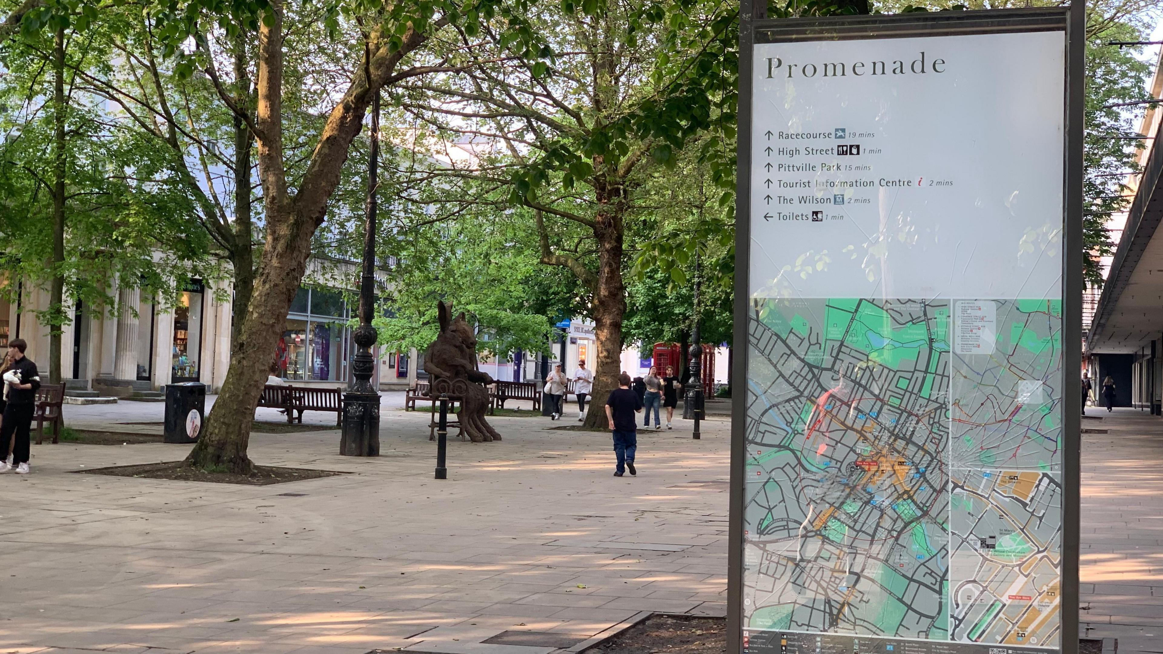 Businesses and shoppers on Cheltenham's tree-lined Promenade. In the foreground there is a noticeboard with a map of the local area. People are walking by - some are carrying bags, others are talking on phones or chatting to friends.