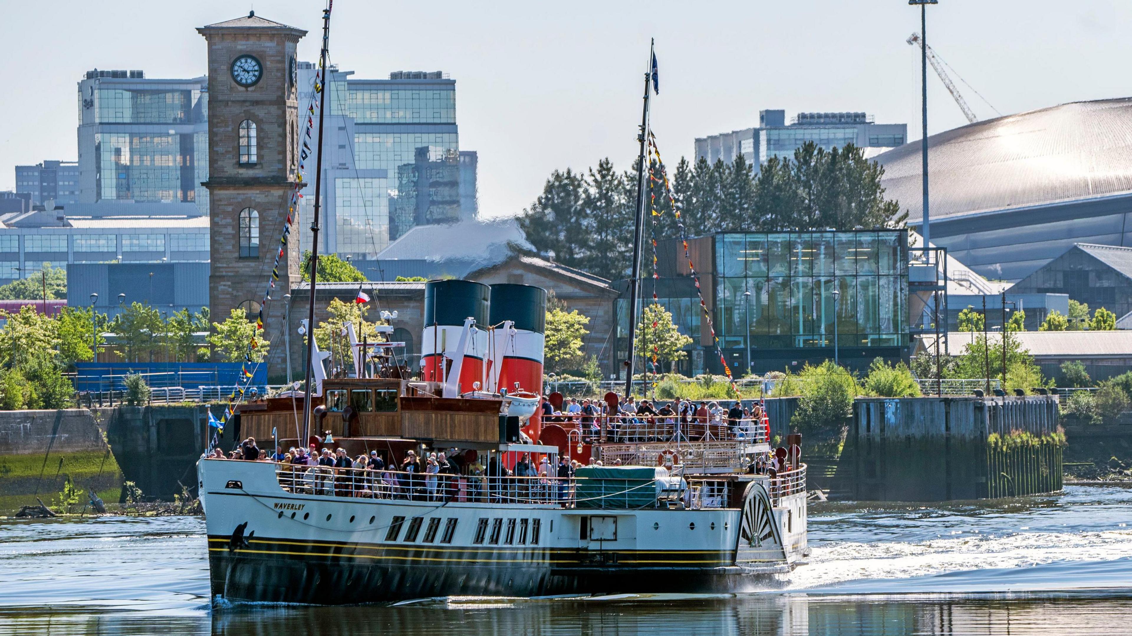 The Waverley sailing near a harbour that has a lot of industrial buildings and a tower with a clock. The vessel has lots of miniature multicoloured flags and passengers on board. 