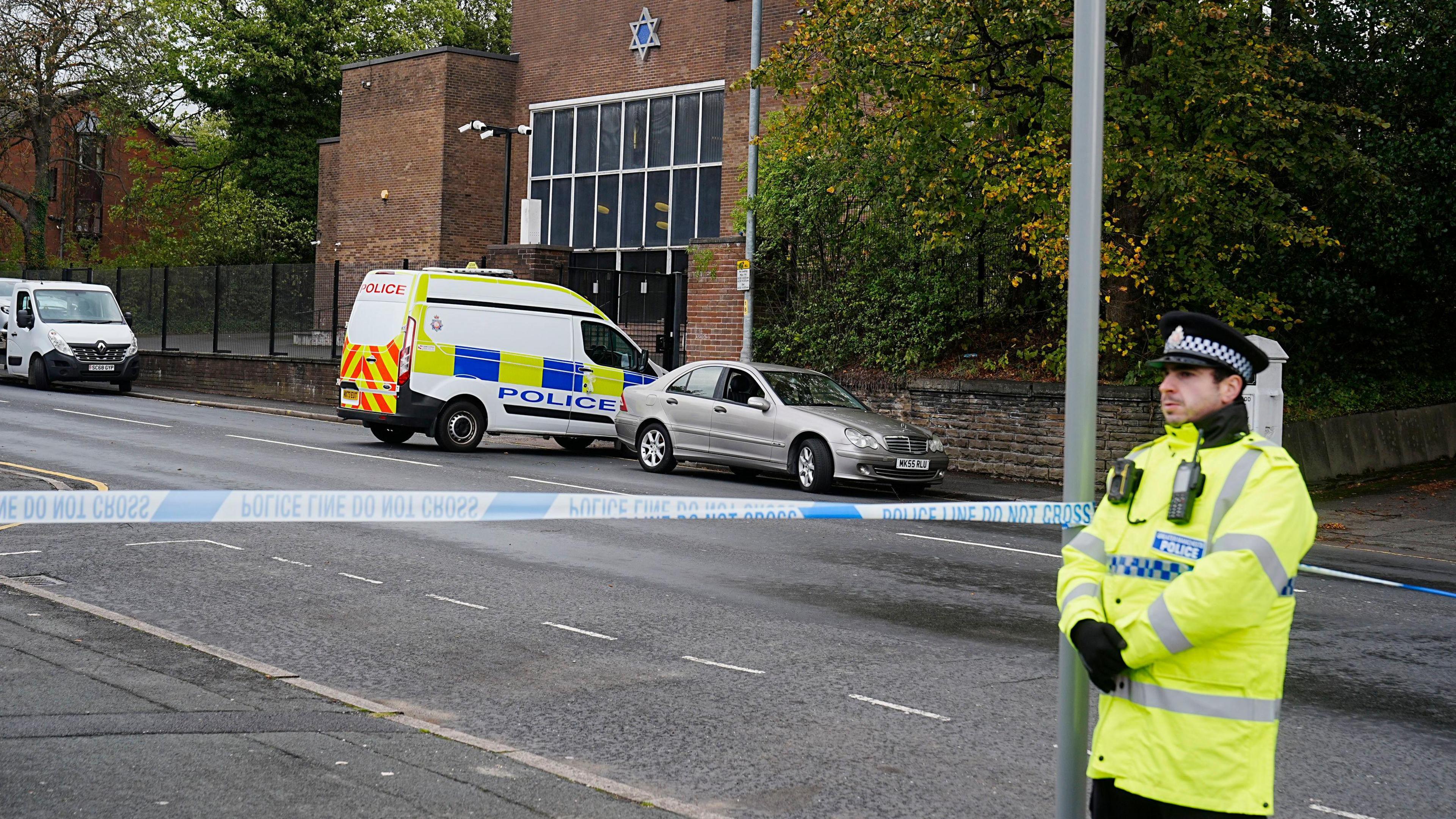 A police officer in a yellow reflective jacket stands by a cordon, across the road from a synagogue. 