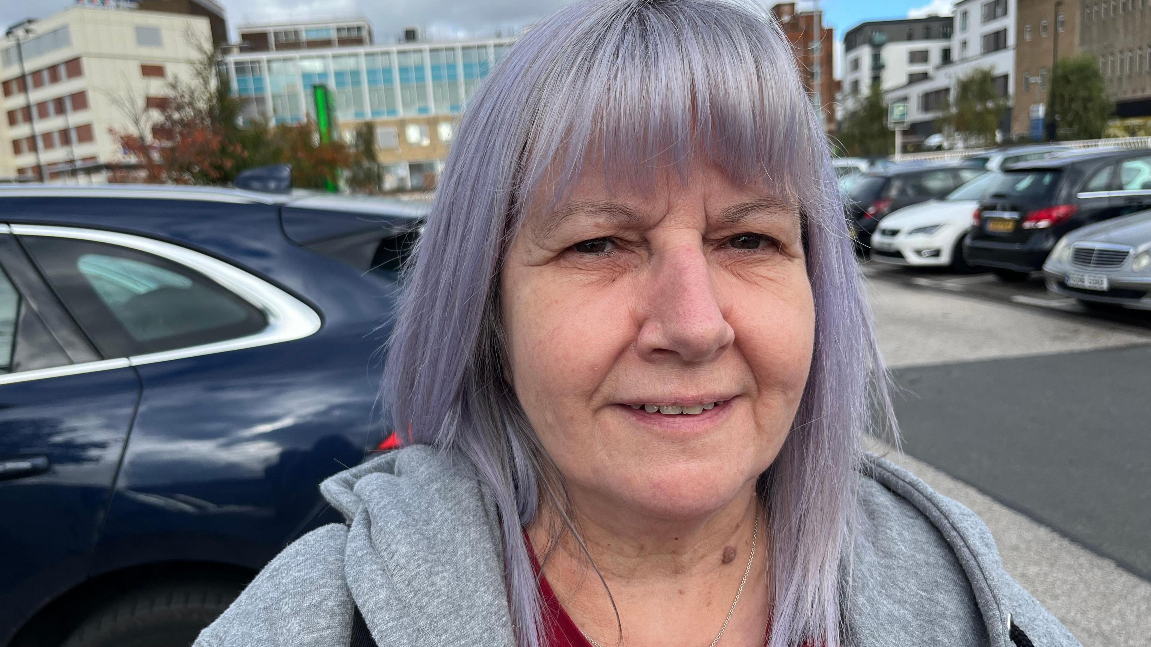 Juliet Wilmore smiling for the camera while stood in a car park. She has long hair that is dyed purple and she is wearing a grey zipped hoodie.