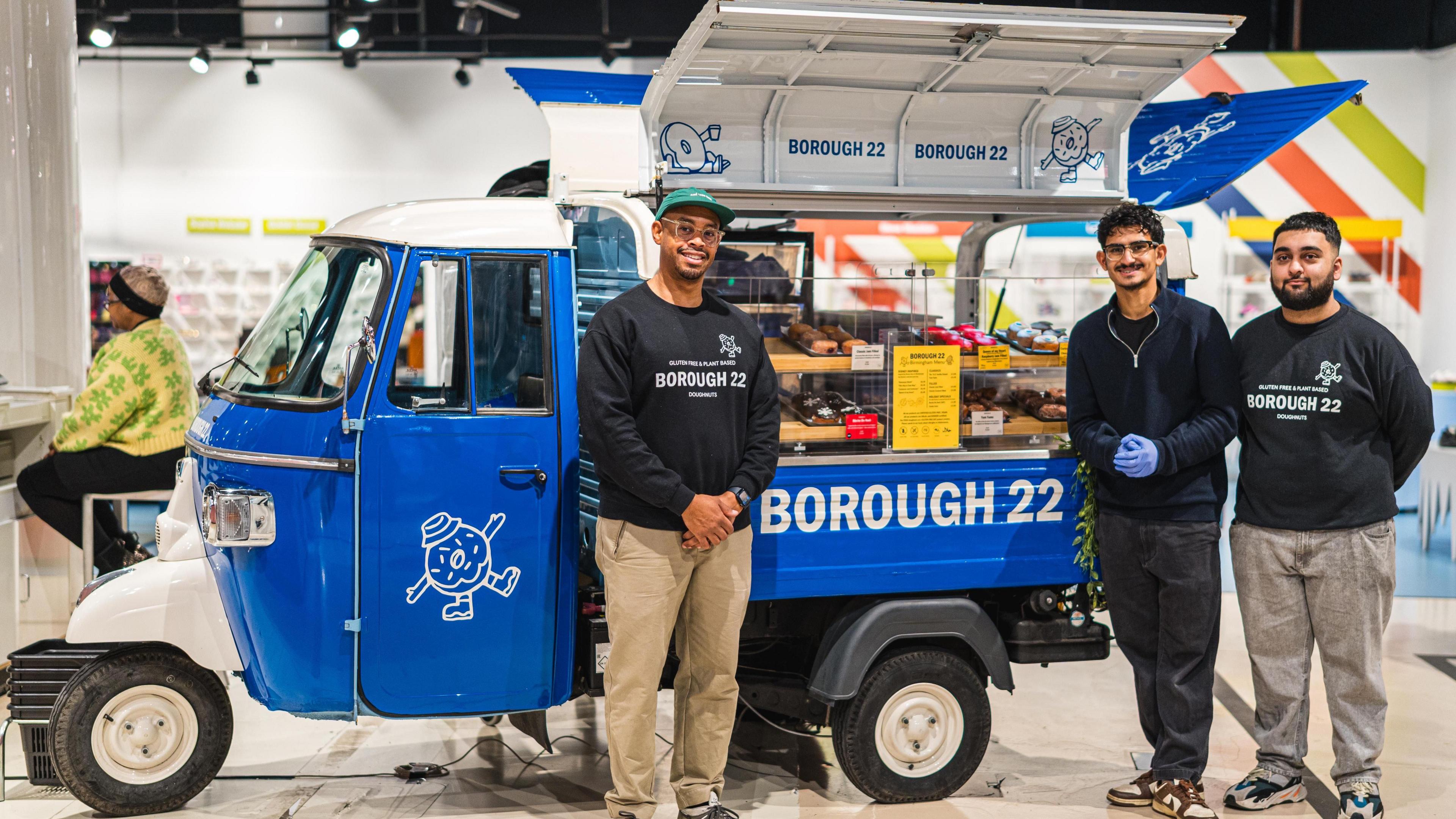 Three men stand in front of a blue food truck in Selfridges which says Borough 22 on it in white writing. Several doughnuts of different colours are on display.