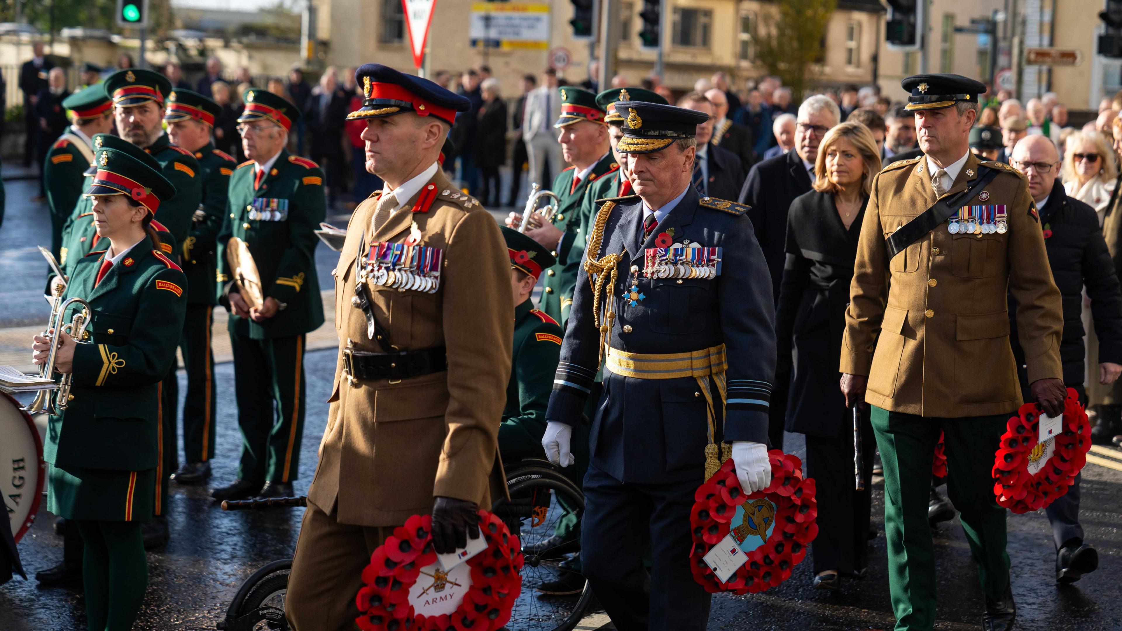 People dressed in army uniforms are marking on a road. Some are holding wreaths and some are holding trumpets. 